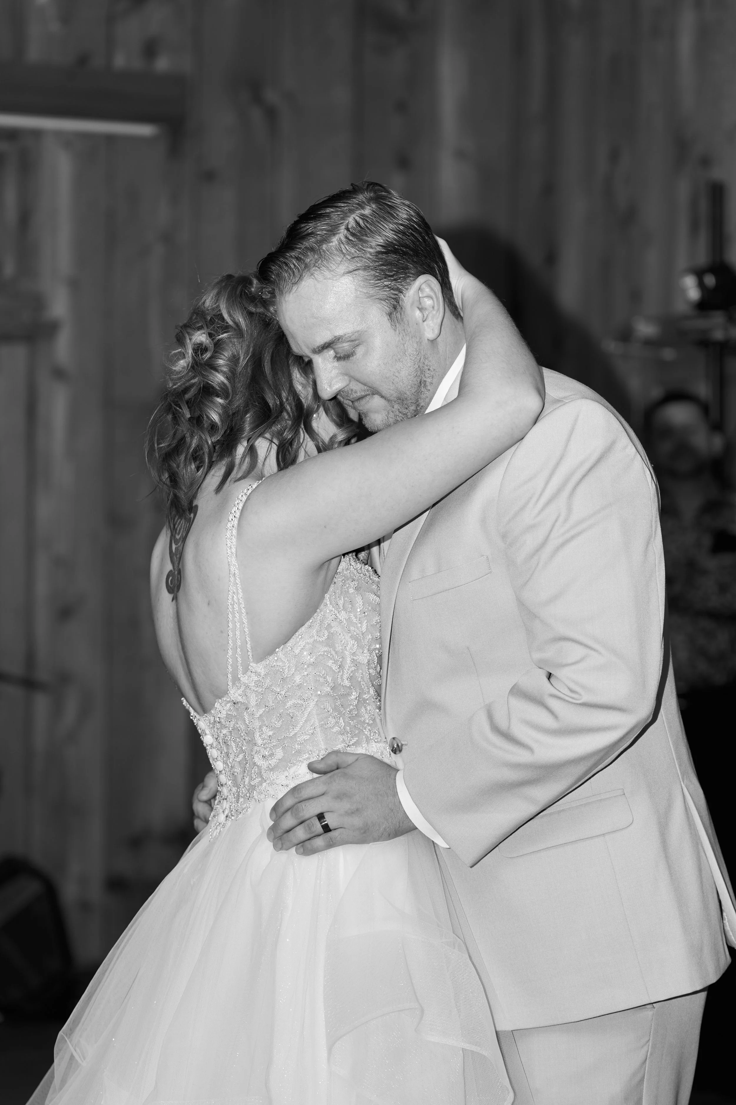 A black and white photo of a couple dancing closely, with their heads touching and eyes closed, in formal wedding attire, in a wooden interior setting.