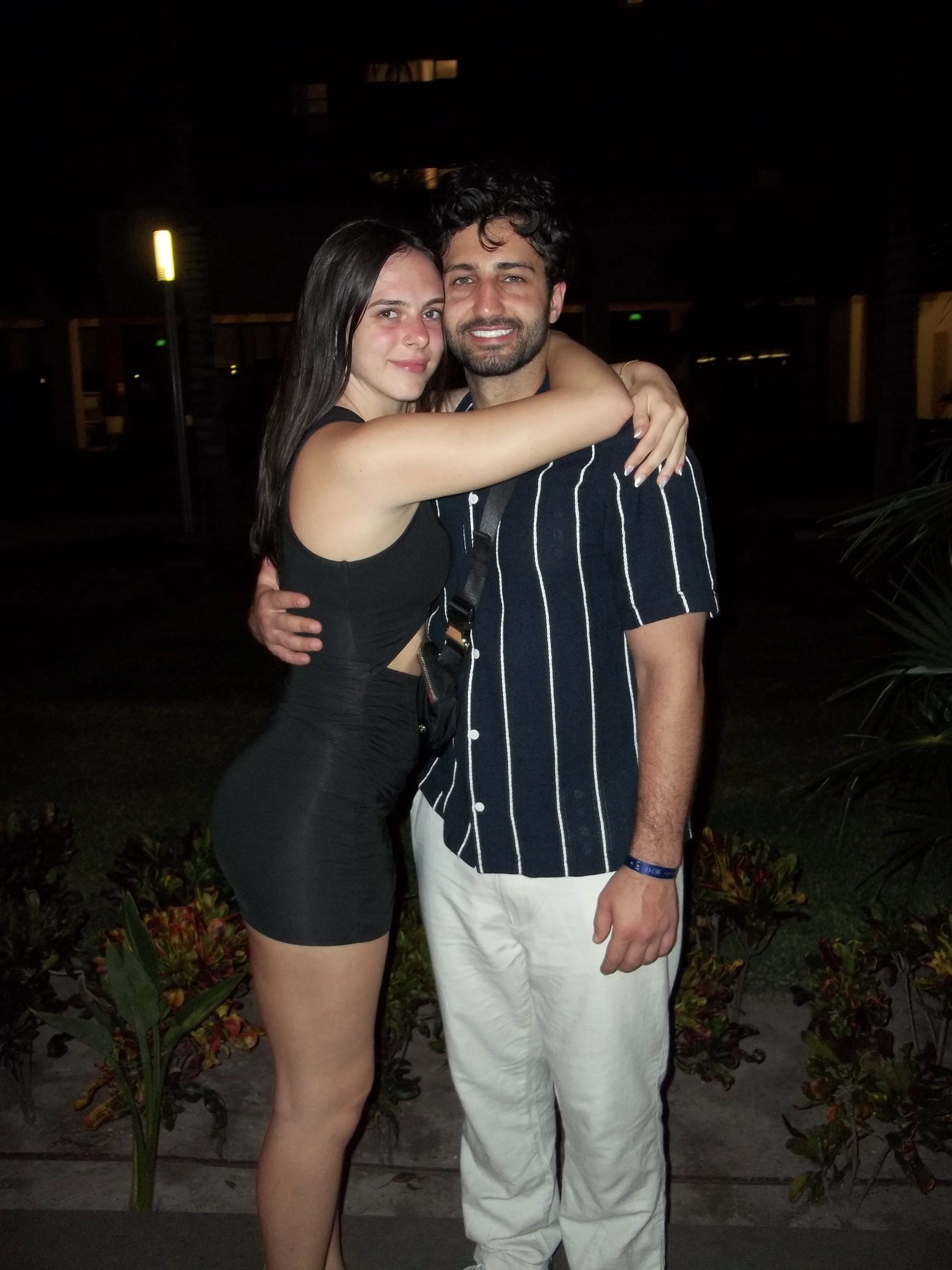 A young woman and a young man hugging at night outdoors. The woman is wearing a black sleeveless dress, and the man is wearing a navy blue striped shirt and white pants.