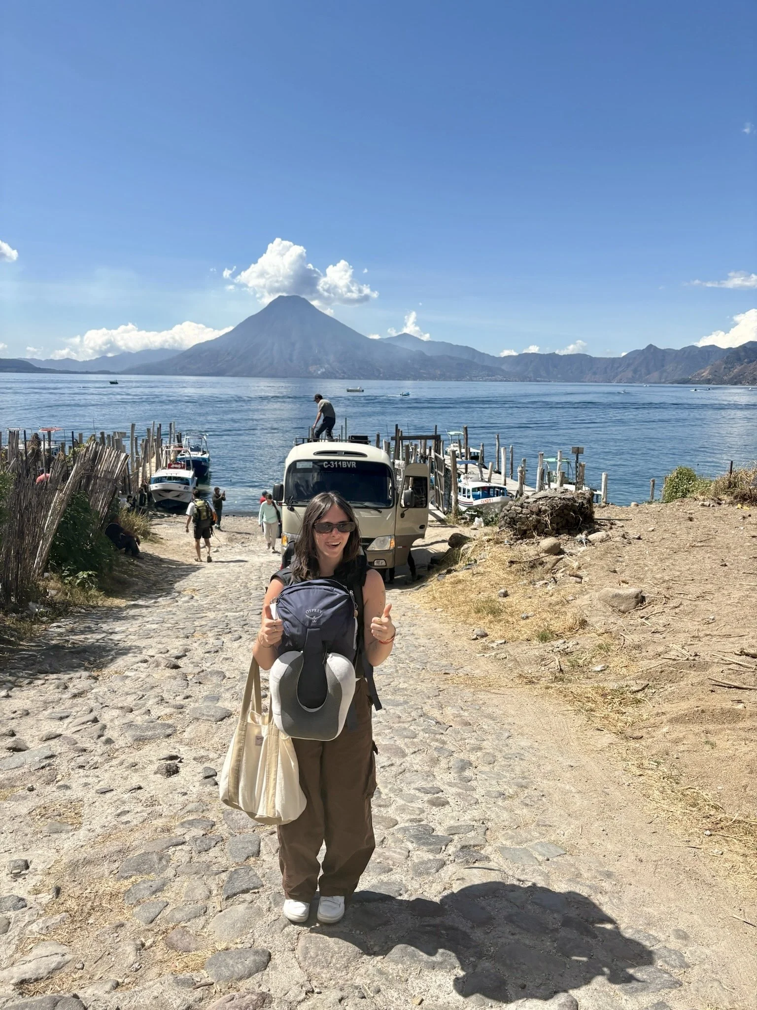 A woman standing on a cobblestone path near a lake, with boats and a dock behind her, and a mountain with a volcano in the distance under a blue sky with clouds.