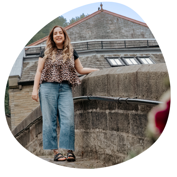 Young woman with long, wavy hair smiling, wearing a leopard print top, blue jeans, and sandals, standing outdoors on a stone pathway in front of a stone building with a red-tiled roof.