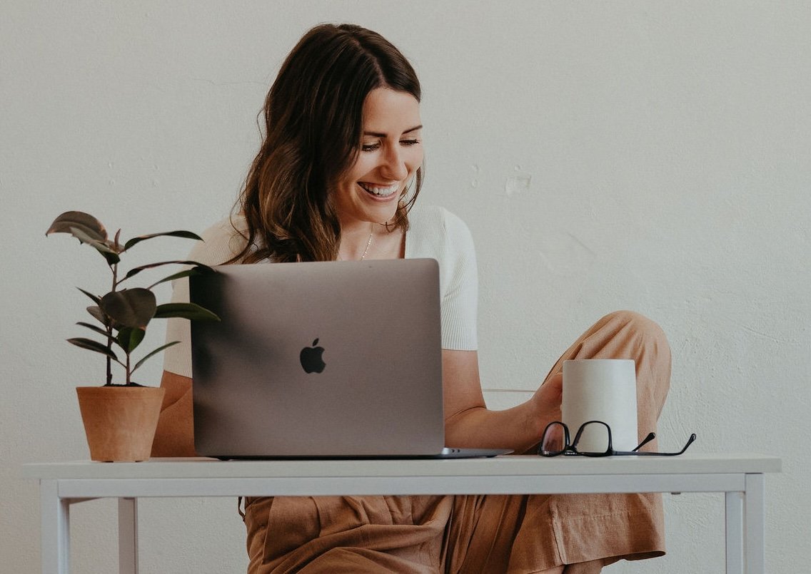 A woman sitting at a white desk with a laptop, a potted plant, a pair of glasses, and a mug, smiling while looking at her phone.