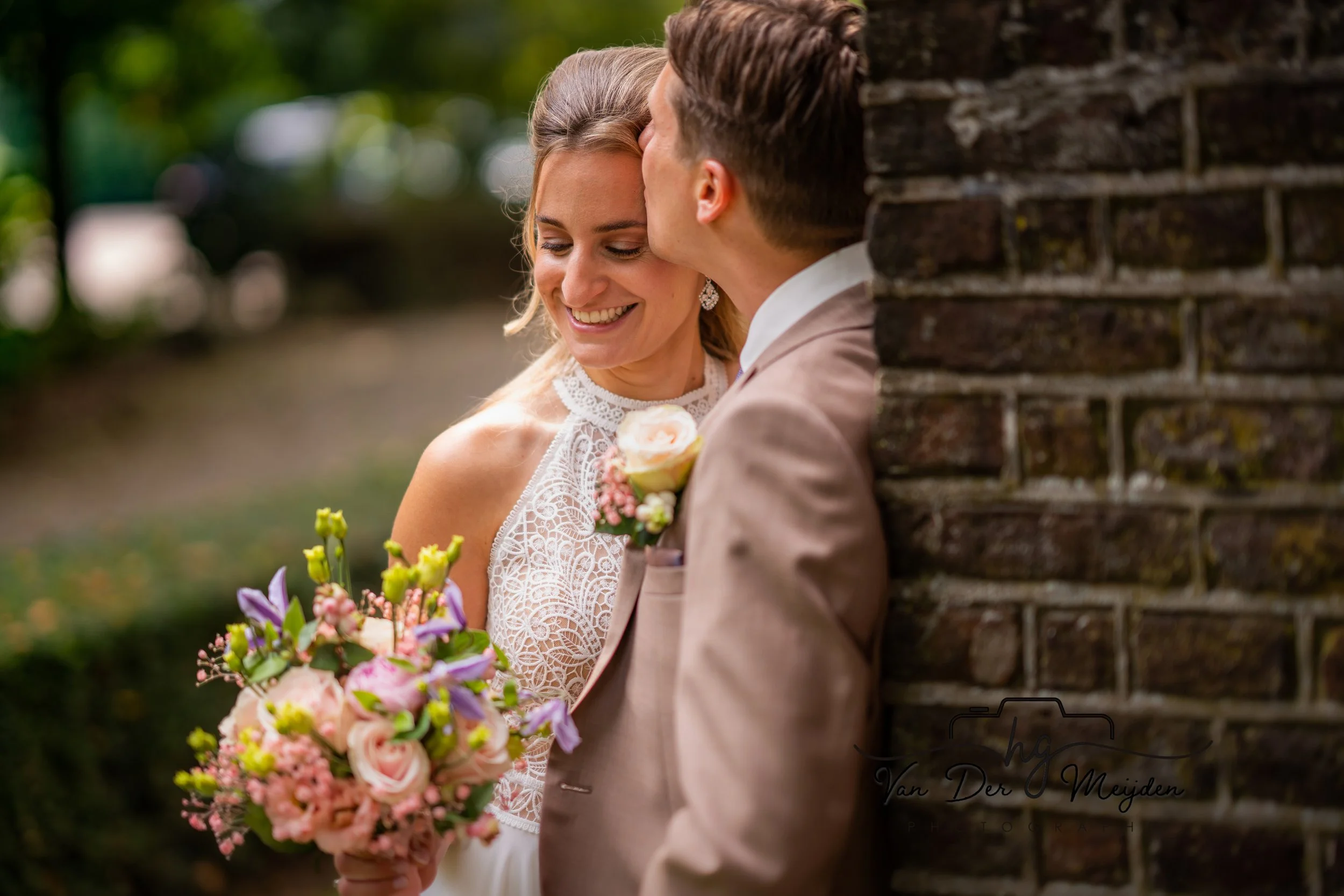 Bruidspaar lachend omarmt elkaar bij bakstenen muur, met bruid in witte jurk en bruidegom in lichtbruine jas, haar boeket vol kleurrijke bloemen houdt ze vast.