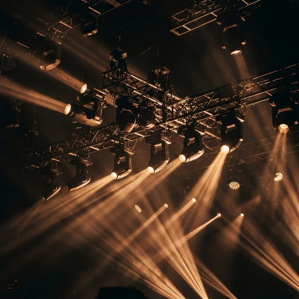 Stage lighting and equipment hanging from a rig, with beam lights creating a dramatic effect in a dark theater or concert venue.