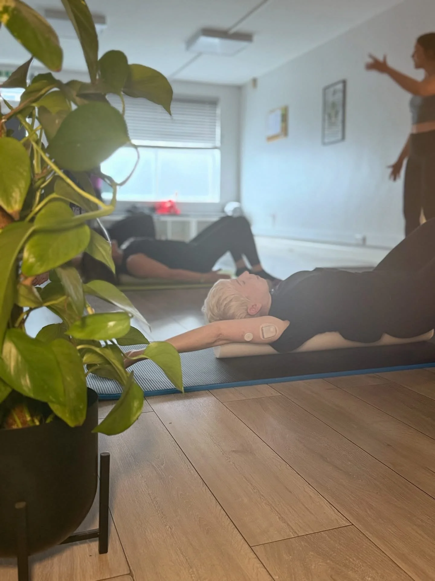 A group of people practicing yoga or fitness exercises on mats in a bright, spacious room, with a woman in the foreground lying on her back with a medical device attached to her arm, and a person standing and gesturing, while another person is in the