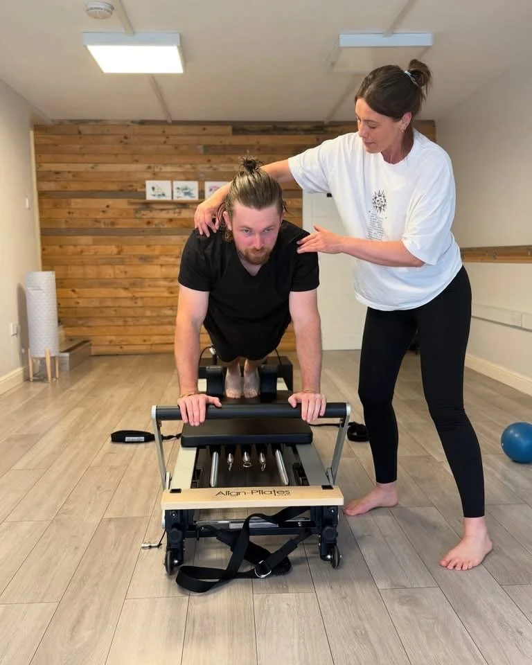 A physical therapist or trainer assisting a man with an exercise on a Pilates reformer machine in a fitness or therapy studio. The man is holding onto the handles in a plank position while the trainer supports him from behind. The room has wooden wal