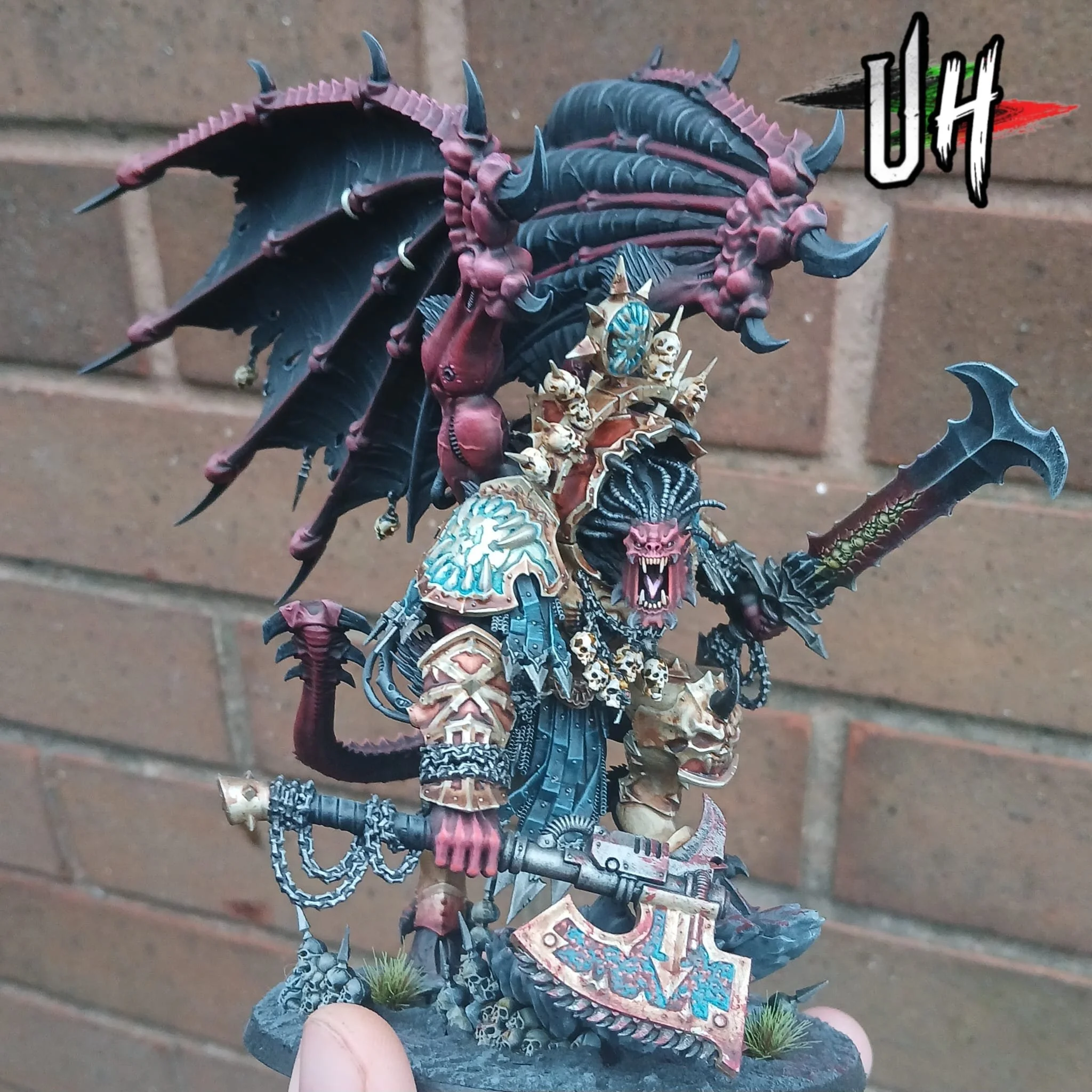 A detailed miniature figure of a fantasy warrior with demonic features, including large dark wings, a lion-like face, skull decorations on armor, and a large axe, set against a brick wall background.