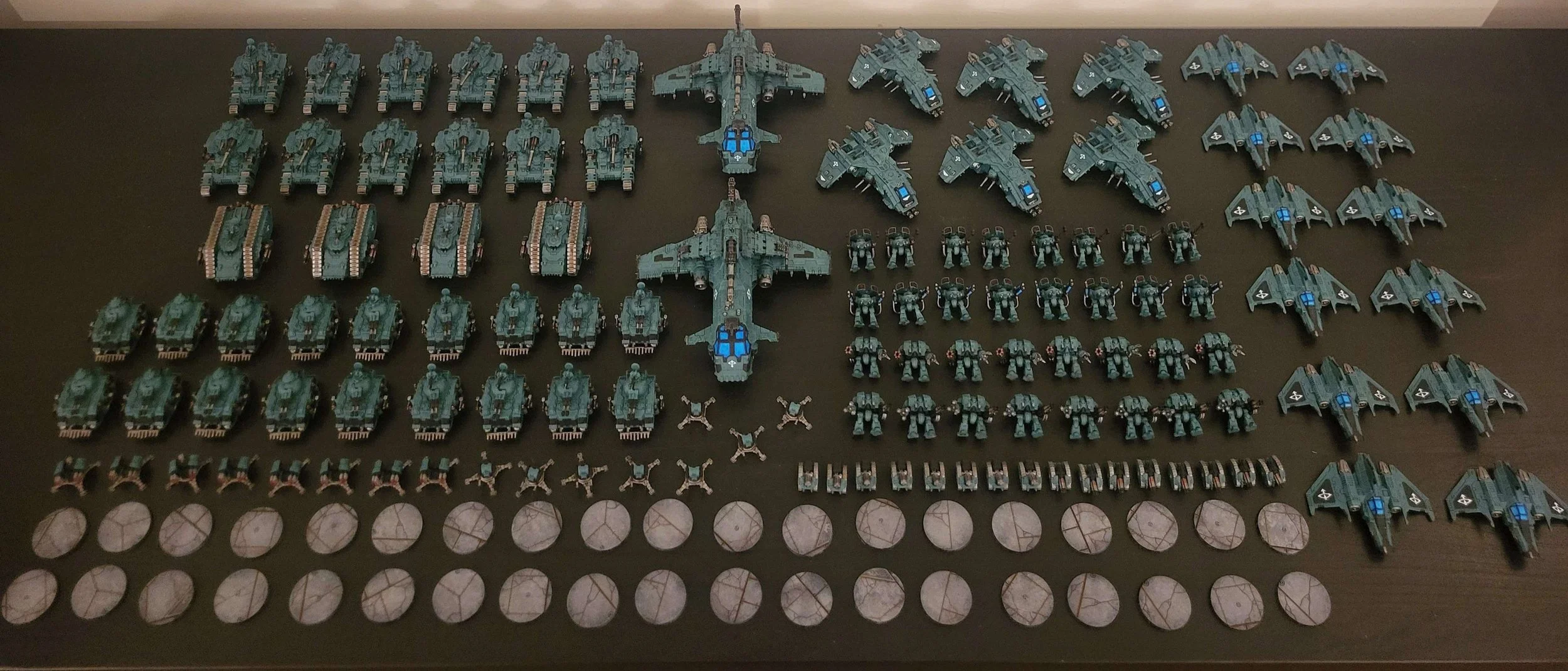 Collection of miniature sci-fi vehicles and figures arranged on a dark surface, including large aircraft, tanks, smaller robots, and round tokens.