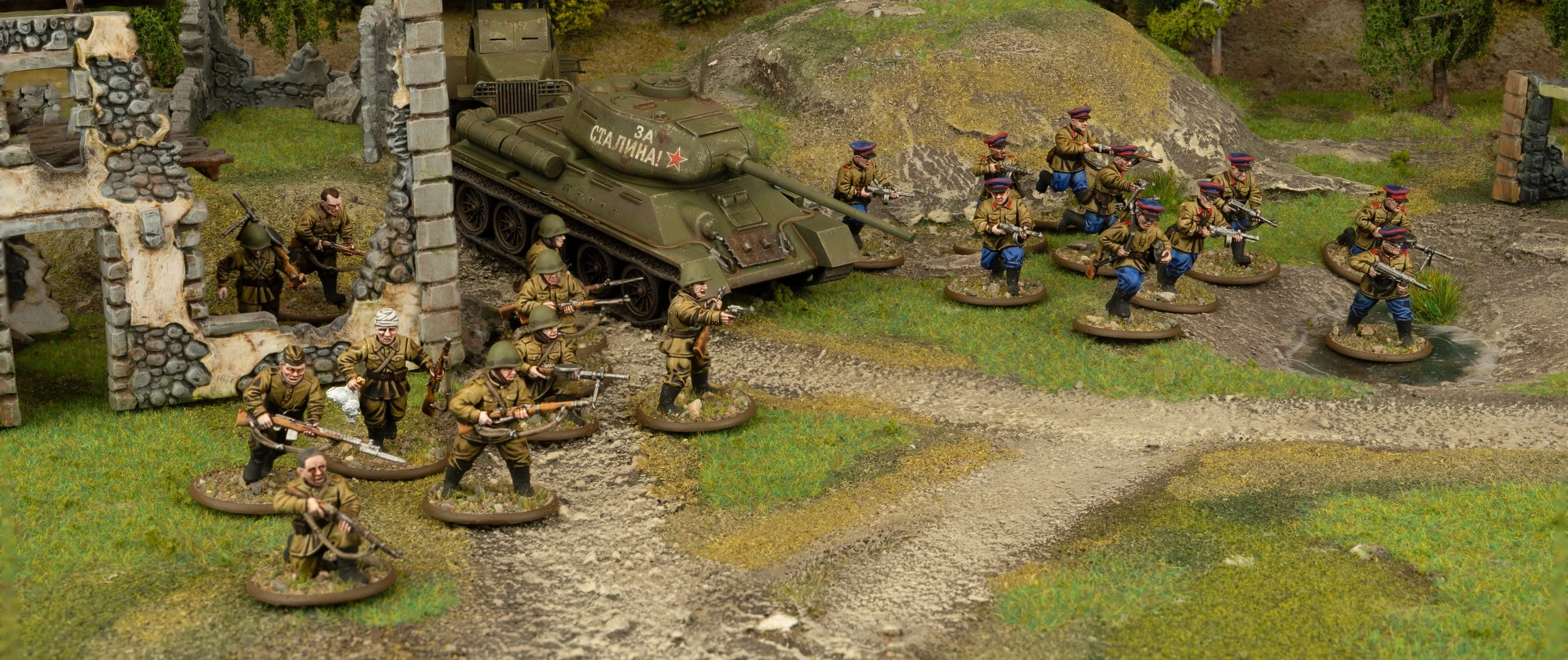 Miniature figures of soldiers with rifles advancing through a wartime scene with ruined buildings and a tank, depicting a military battle diorama.