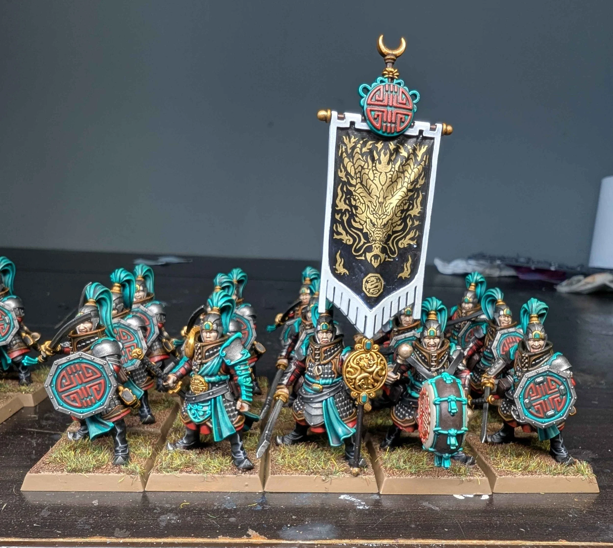 A detailed miniature army of fantasy soldiers with armor, shields, and weapons, featuring a central banner with a dragon decal, painted in black, gold, silver, red, and teal colors, arranged on a display stand with grass-textured bases.
