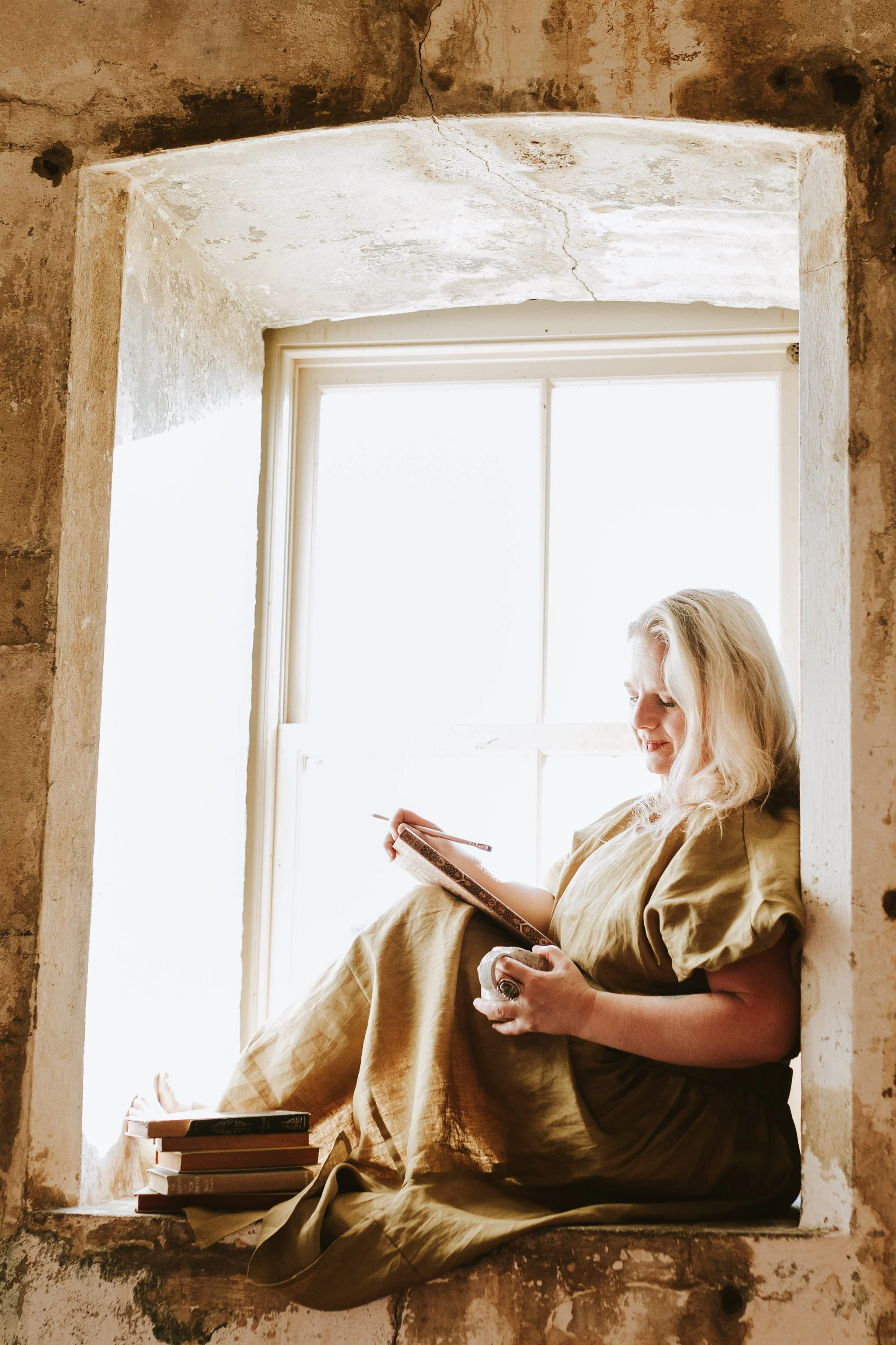 Woman sitting on a windowsill reading a book with a stack of books at her feet, backlit by bright sunlight through the window.