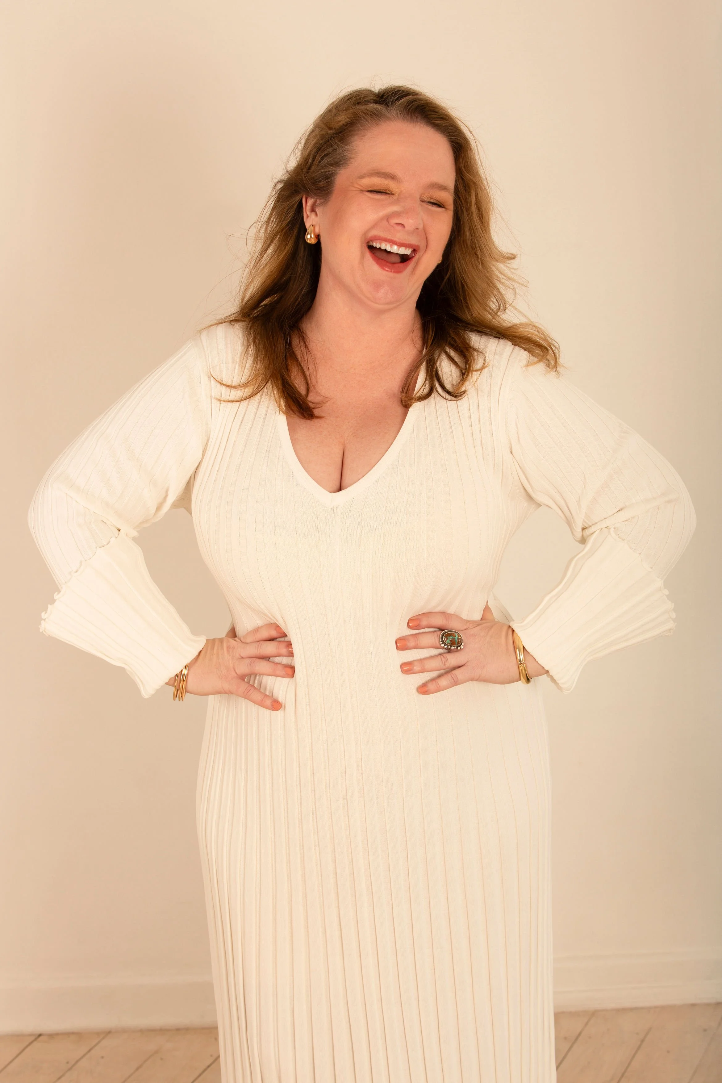 A woman with brown hair is standing indoors against a plain light-colored wall. She is wearing a long, cream-colored, pleated dress with long sleeves and a V-neckline. She is smiling and laughing, with her eyes partially closed, and has her hands on her hips. She is accessorized with gold earrings, multiple gold bracelets, and a large statement ring on her right hand.