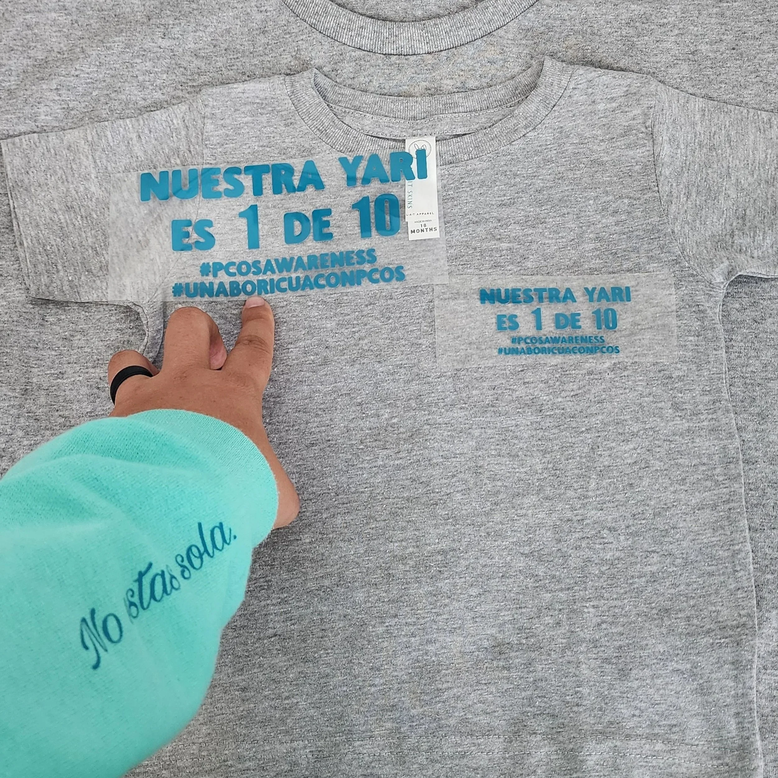 Dos camisetas grises con texto en azul; la camiseta más grande dice 'Nuestra YARI es 1 de 10 #PCOSAWARENESS #UNABORICUAONCOS', y la más pequeña dice también lo mismo en tamaño reducido. Una mano con un anillo en el dedo índice toca la camiseta más gr