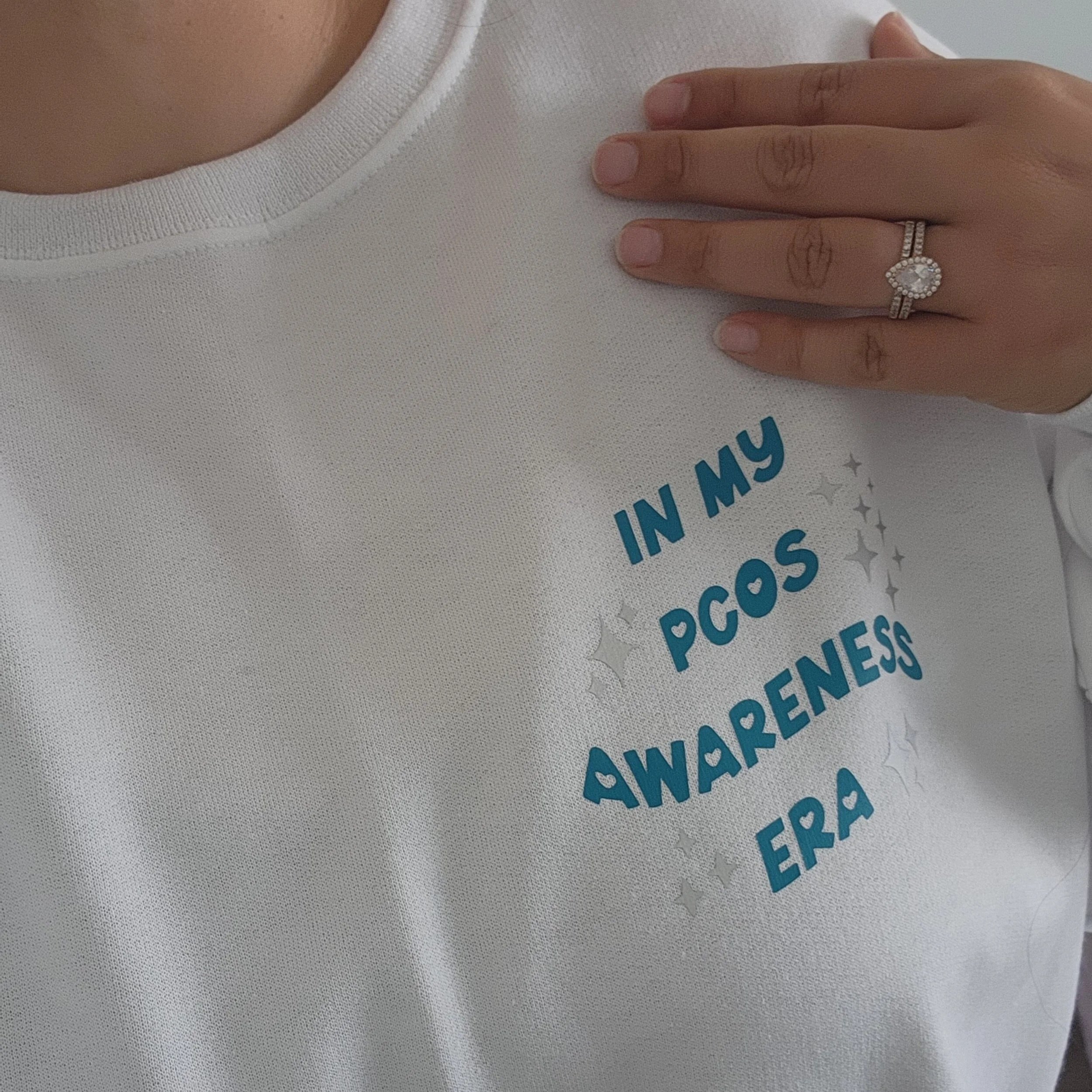Persona usando una camiseta blanca con el texto "In my PCOS awareness era" y adornada con pequeñas estrellas plateadas. La persona lleva un anillo brillante y tiene tatuajes en los dedos.