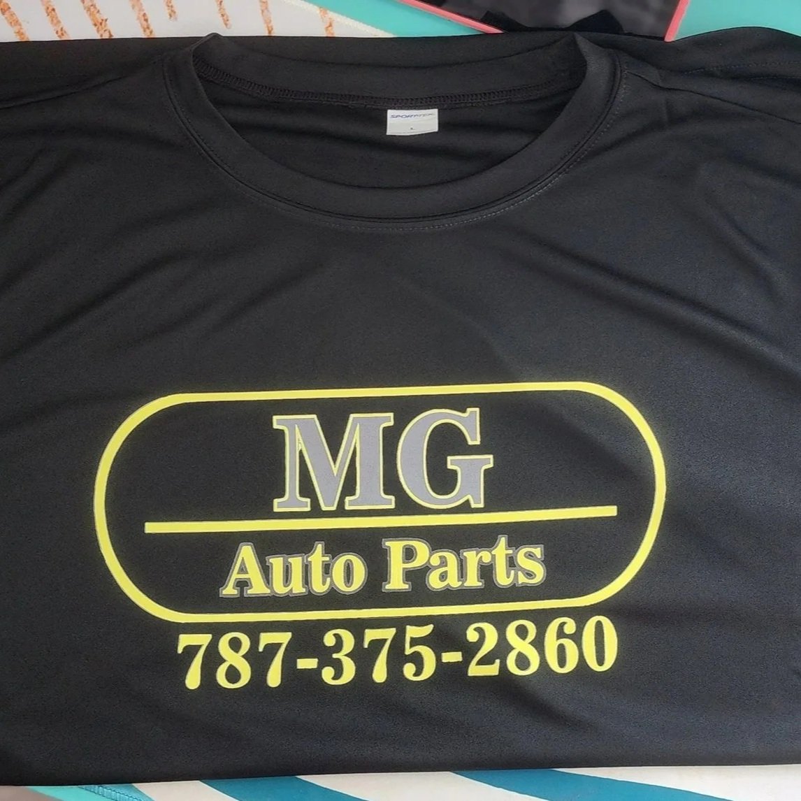 Camiseta negra con logotipo amarillo y gris que dice 'MG Auto Parts' y un número de teléfono debajo.