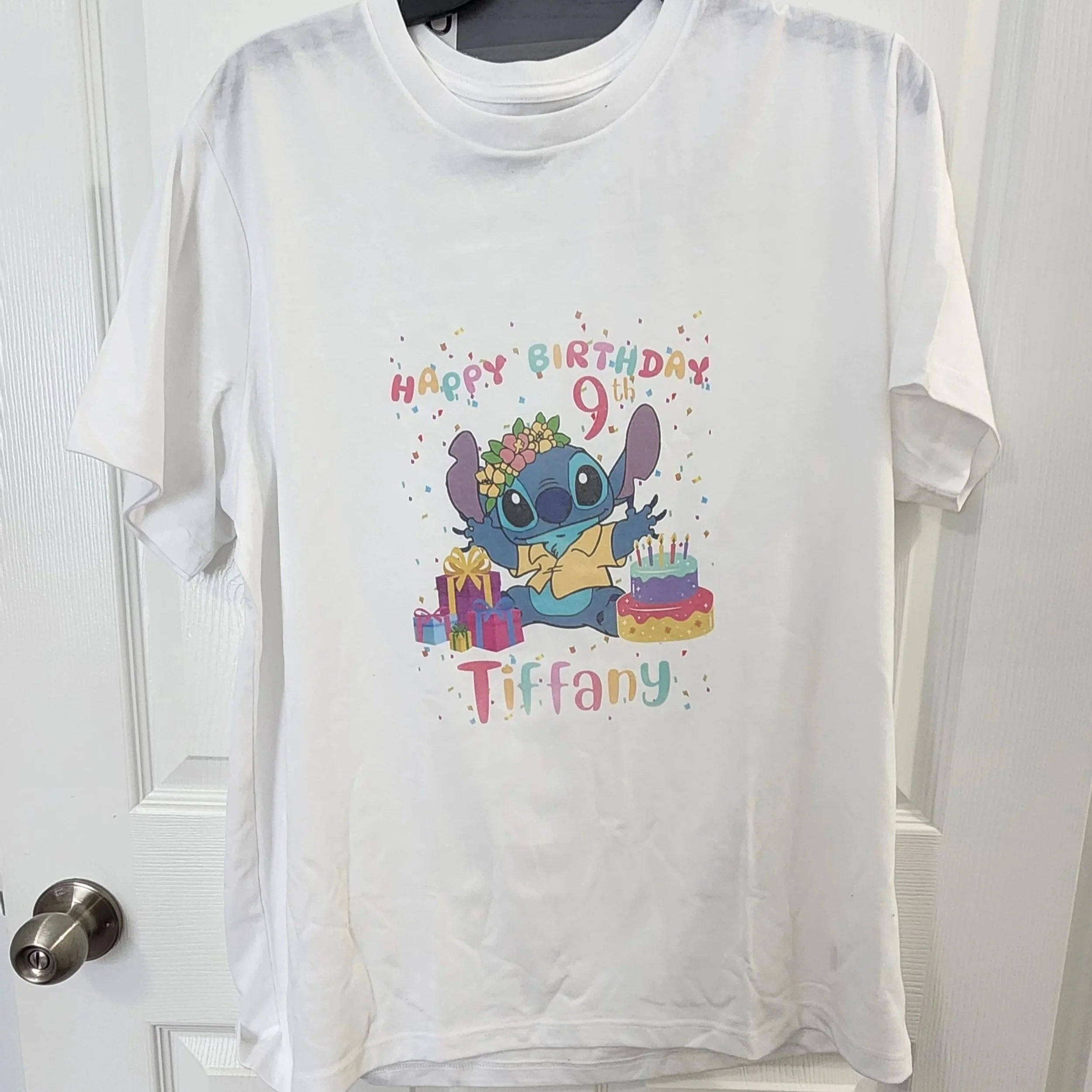 Camiseta blanca con la imagen de 'Stitch' de Disney con corona de flores, rodeado de regalos y un pastel de cumpleaños, con el texto 'Feliz cumpleaños Tiffany' y 'Happy Birthday 9th' en colores pastel.