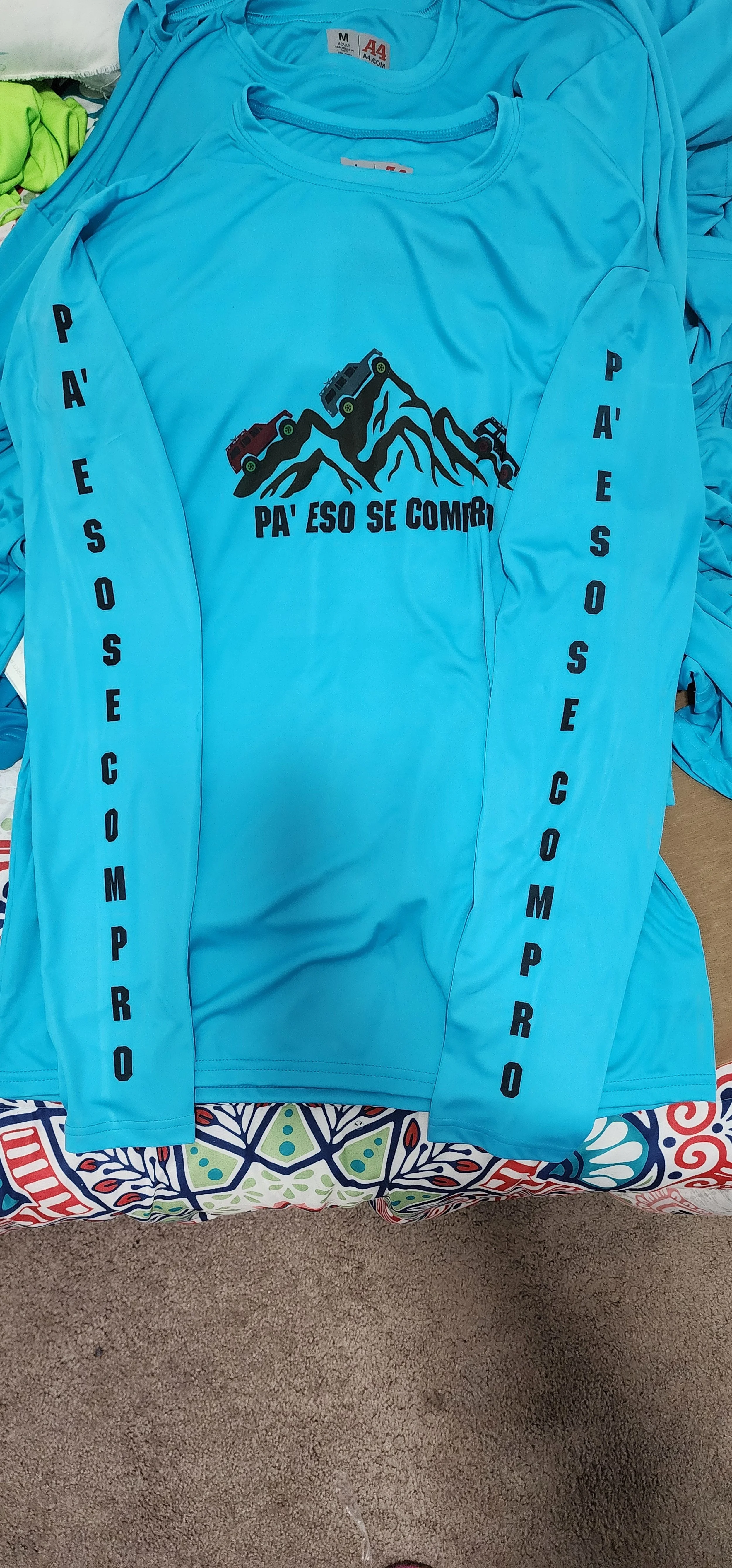 Camiseta azul con una ilustración de montañas y carros en la parte frontal, y las palabras en español 'PA'ESO SE COMPR' en la parte central, y 'PA'ESO SE COMPR' en las mangas.