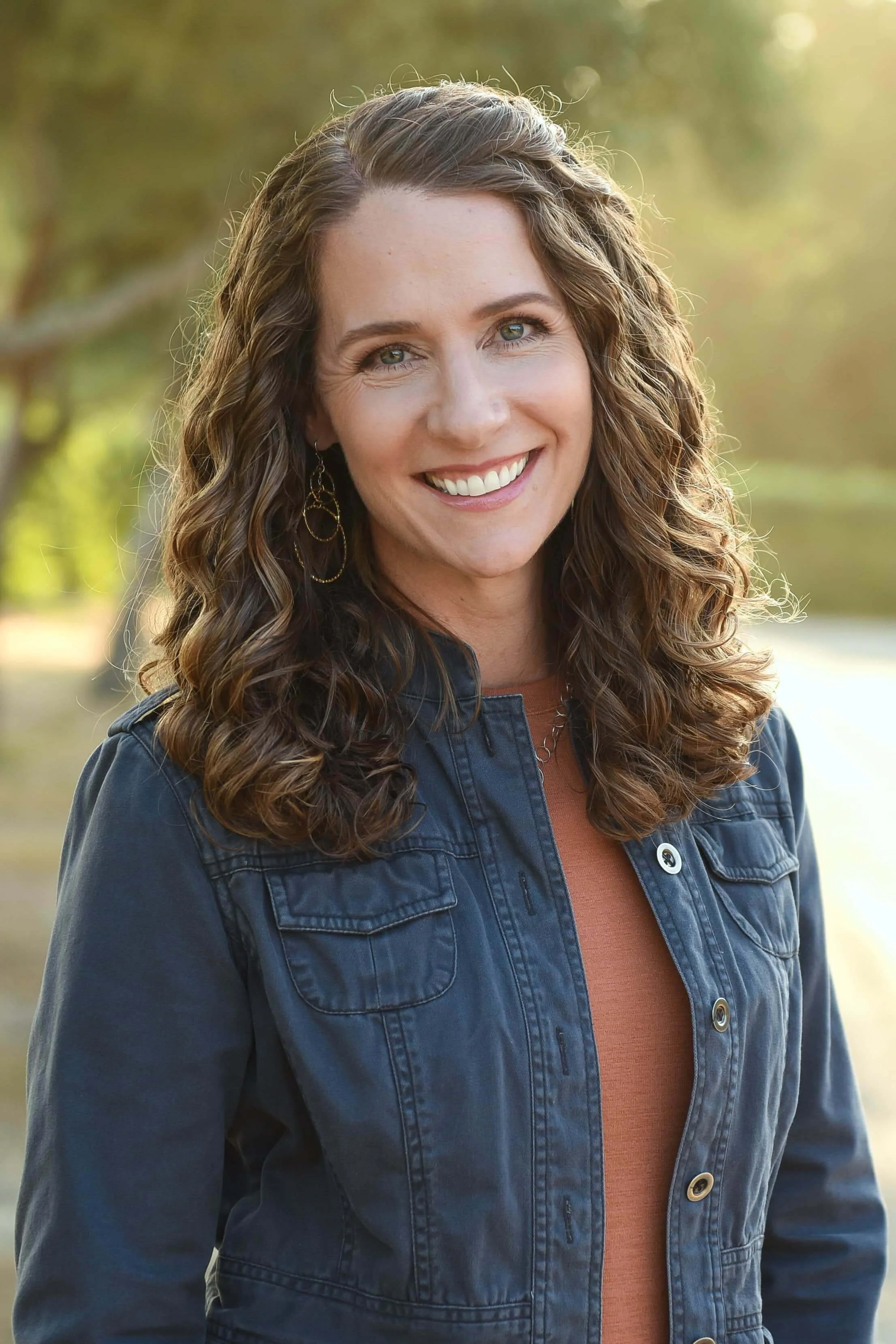Woman with curly hair smiling outdoors in a denim jacket, wearing hoop earrings, and standing in front of a blurred natural background.