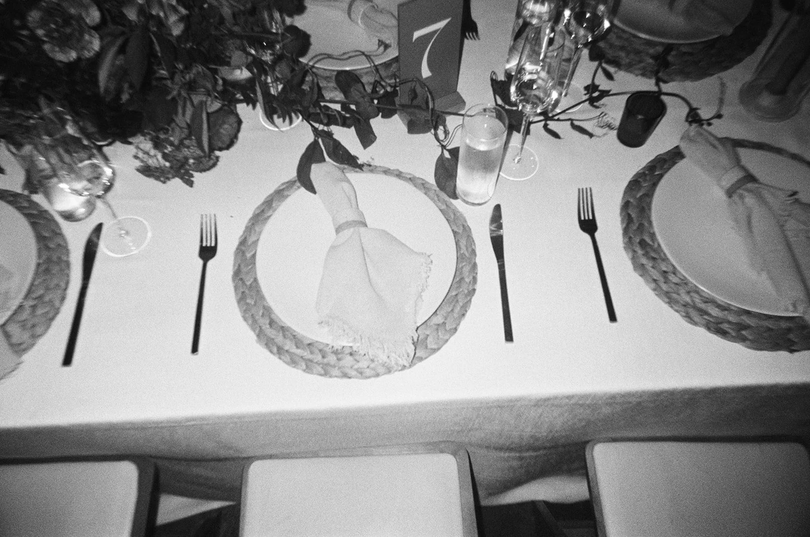 Dining table setting with plates, silverware, a folded napkin, wine glasses, water glasses, and a centerpiece with foliage and a table number sign with the number 7.
