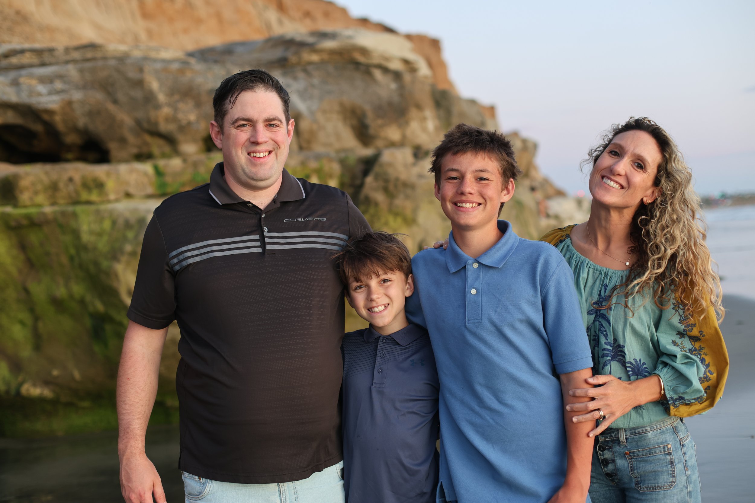 Family of four smiling on the beach with cliffs and ocean in the background.