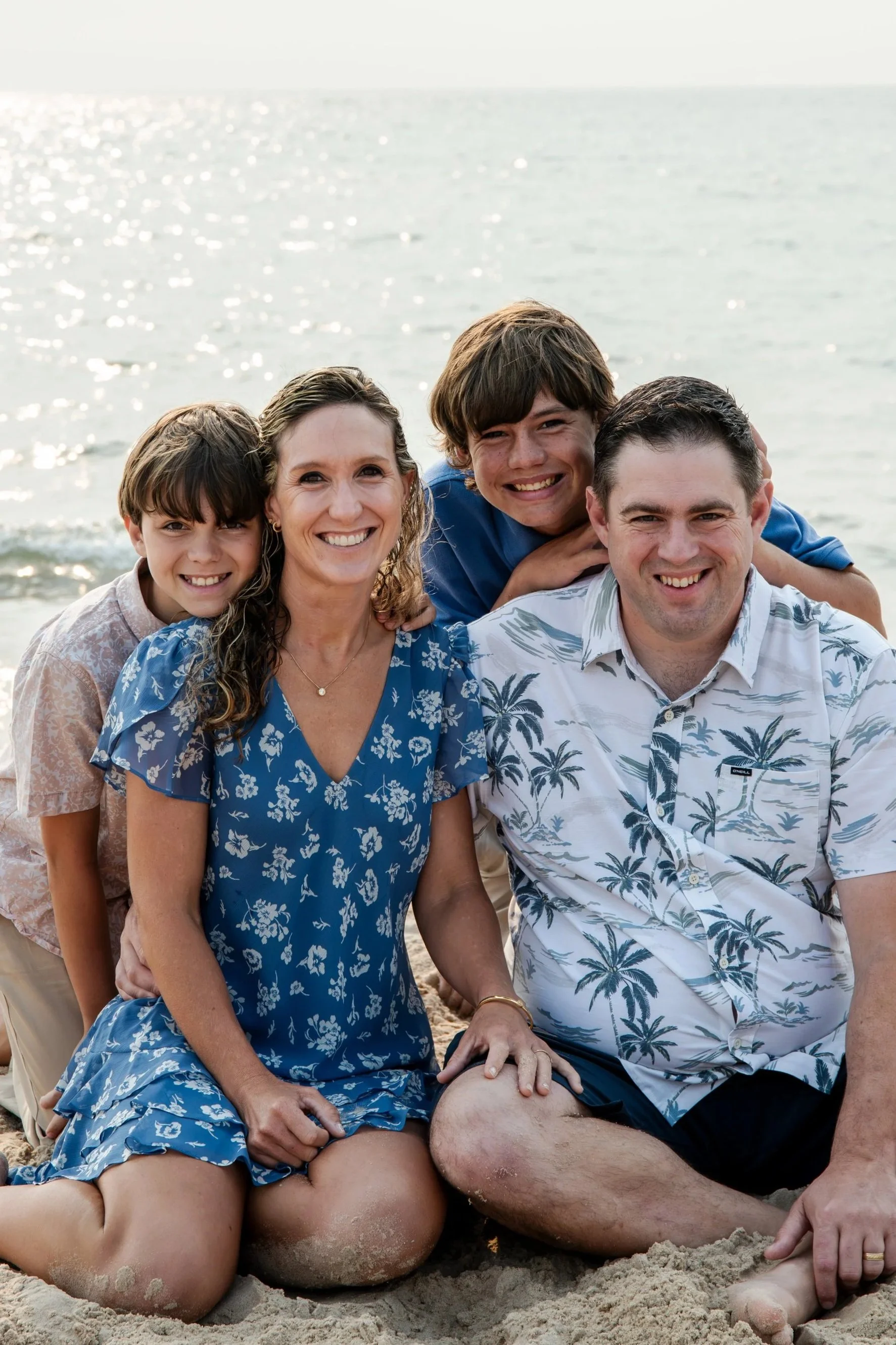 Family of four sitting closely together in the sand, smiling at the camera with two boys leaning over their parents.