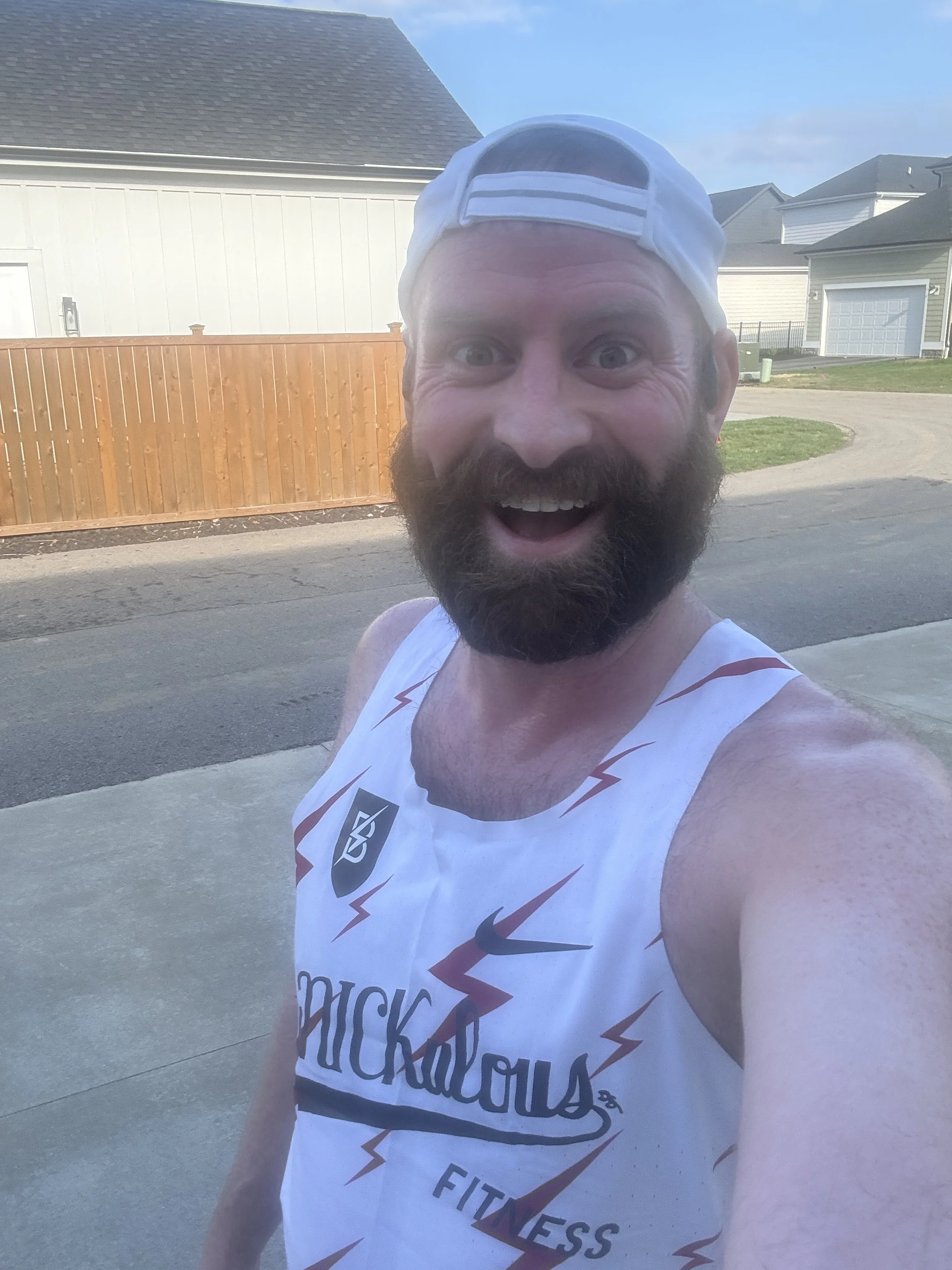 A smiling bearded man wearing a white RiNickulous tank top and a white backward cap, taking a selfie outdoors in a residential neighborhood with houses, a wooden fence, and a clear blue sky in the background.
