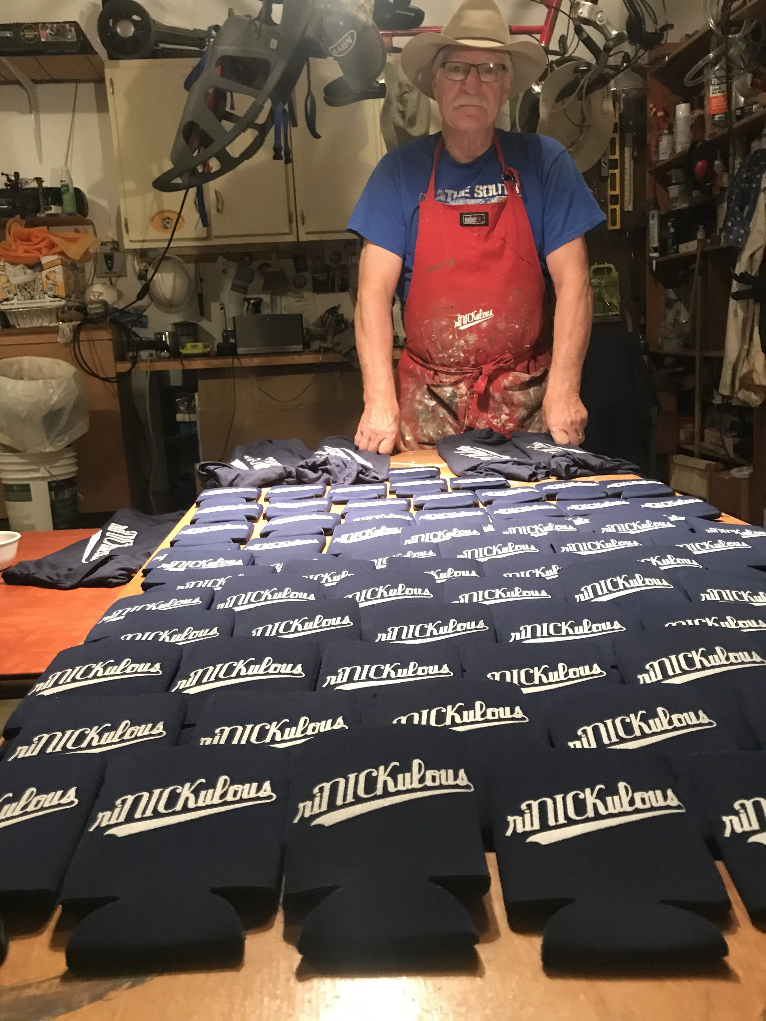 A man wearing a cowboy hat, glasses, a blue t-shirt, and a red apron standing behind a table with multiple printed 'RiNickulous' in T-Shirt's. The background shows a cluttered workspace with shelves, bicycles, and various tools.
