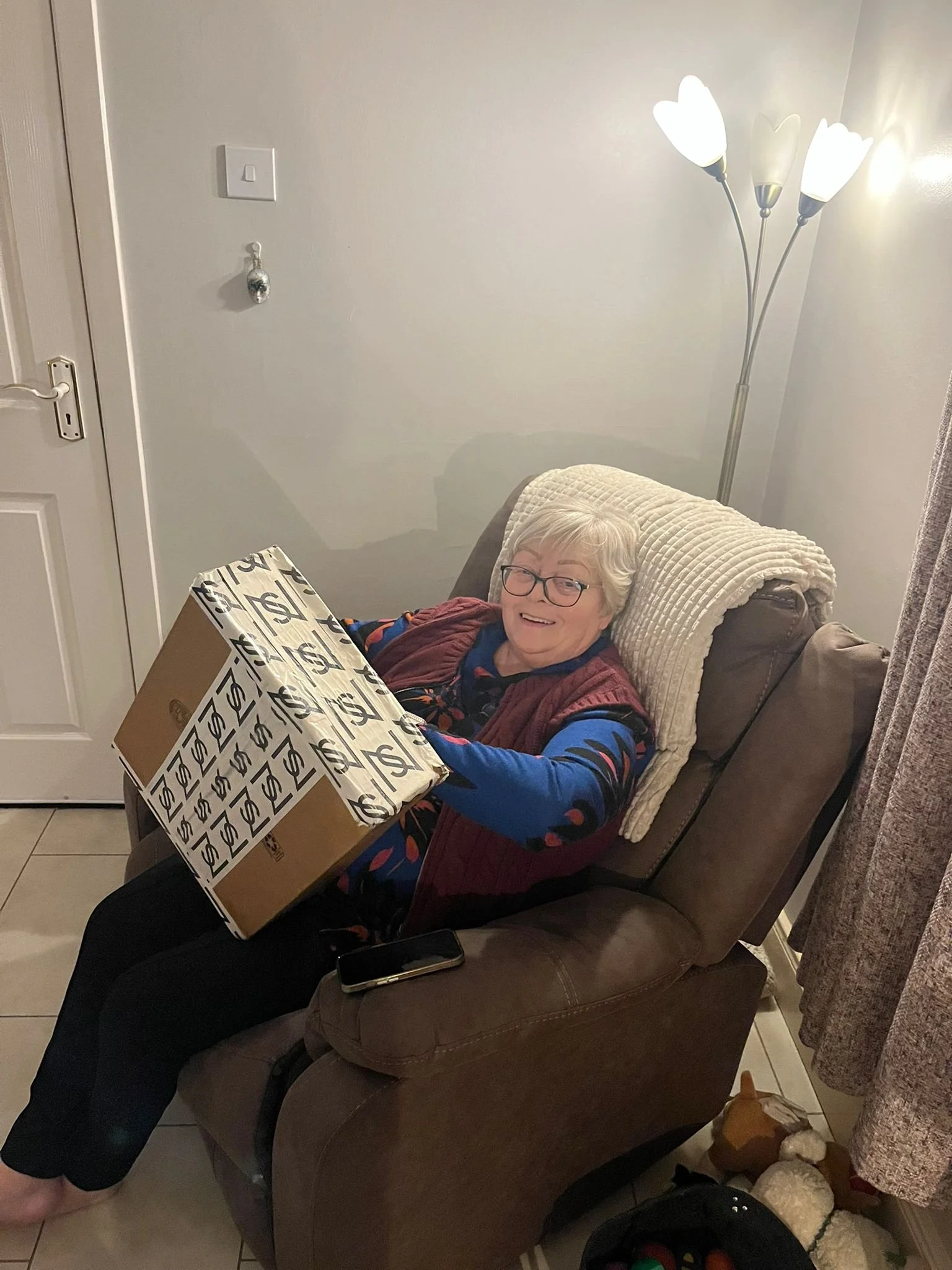 An elderly woman with glasses smiling while sitting on a brown recliner chair and holding a large gift box wrapped with white paper featuring black dollar signs. She is indoors, with a cream blanket on her shoulders and surrounded by various items.