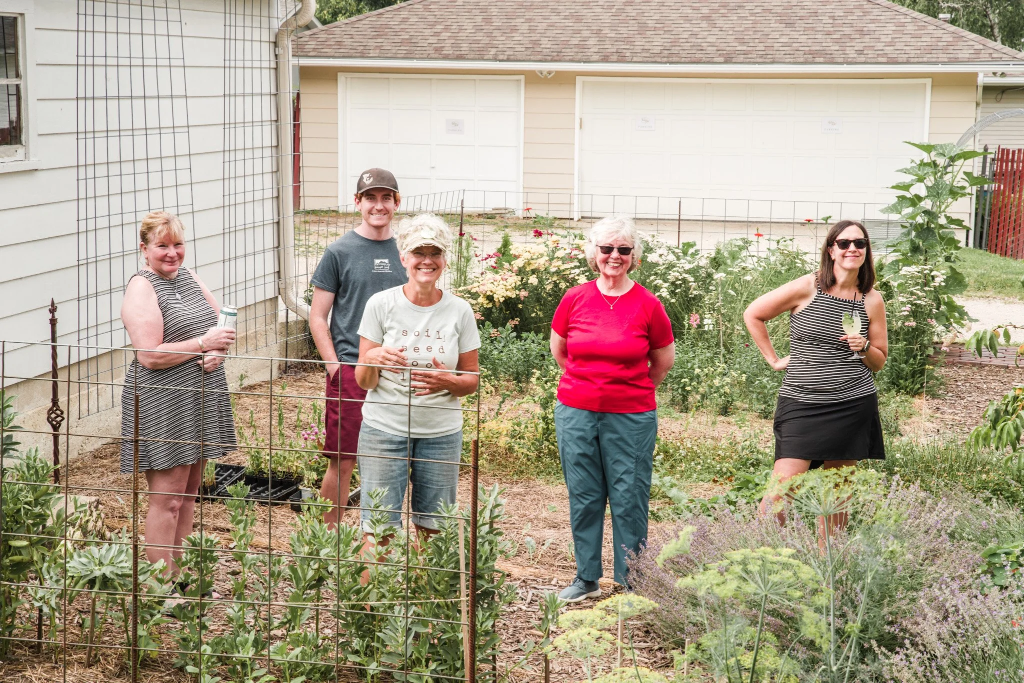 Group of five women and one man standing behind a garden fence, smiling, in a garden with flowers and plants, in front of a white house with garages and trees in the background.