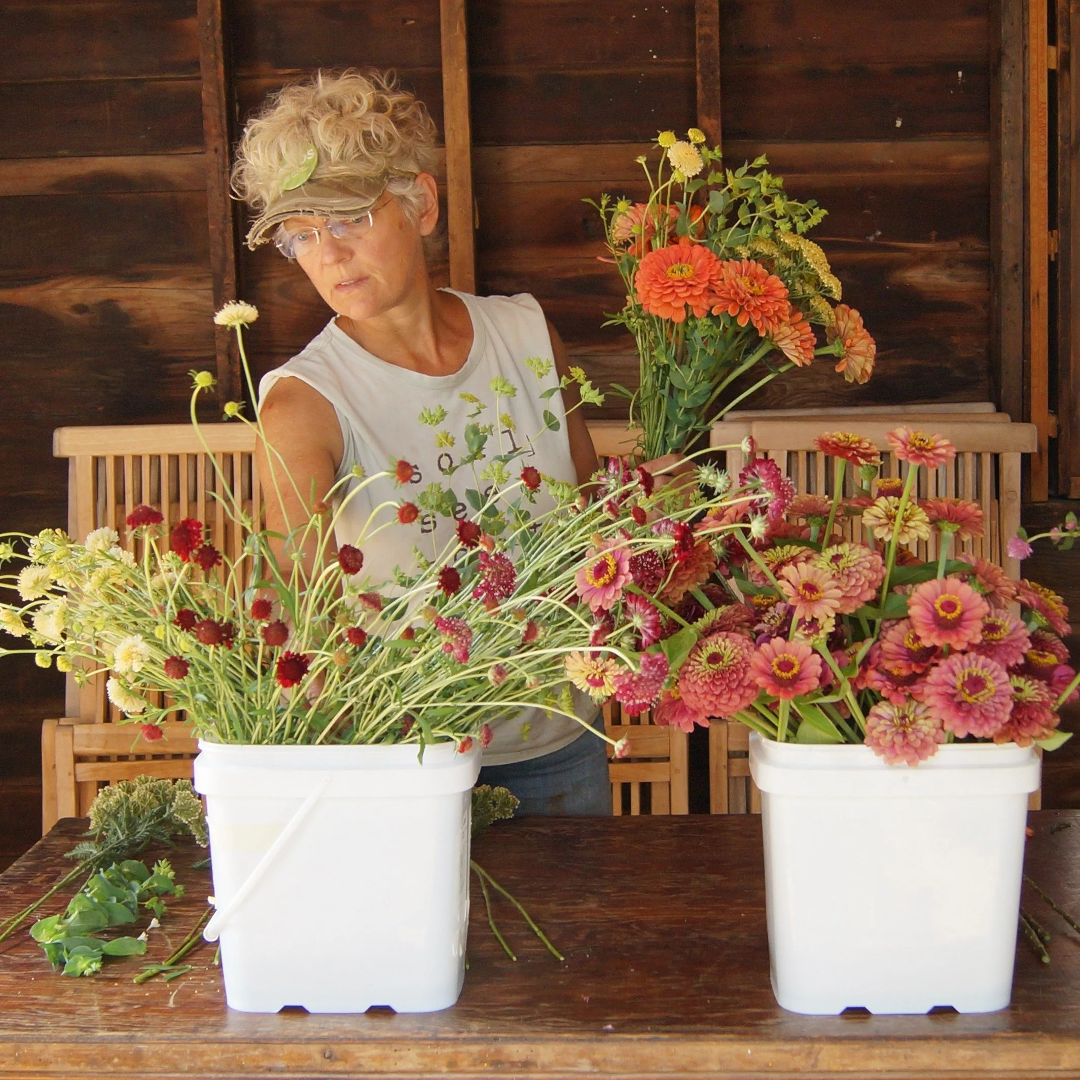 Various colorful flowers in white containers on a wooden table, with a woman in a white shirt arranging the flowers, dark wooden wall in the background.