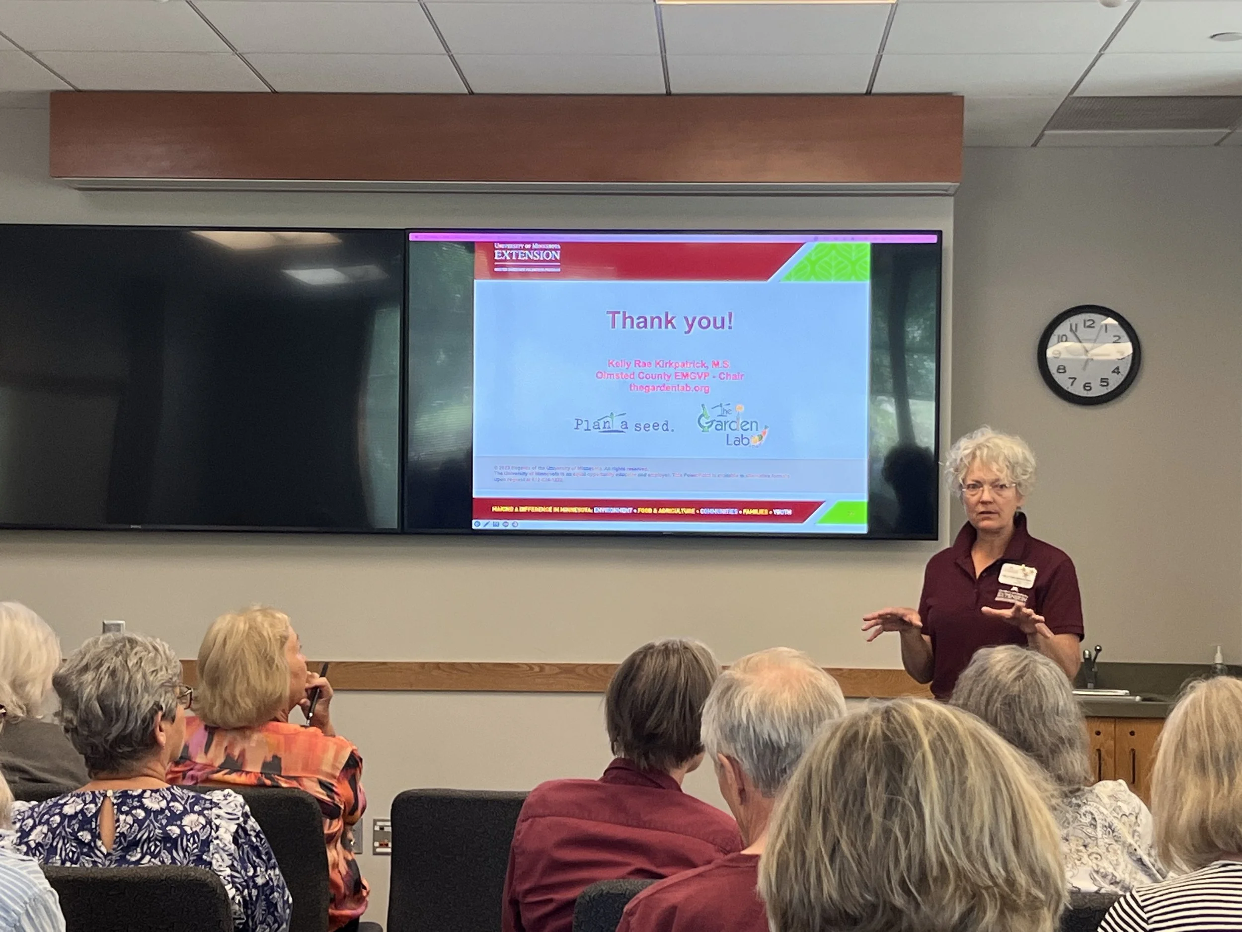 A group of older adults seated in a room, listening to a woman giving a presentation. A large screen behind her displays a thank you message from the University of Minnesota Extension, including a website and sponsors. The room has a wall clock showing 9:14.