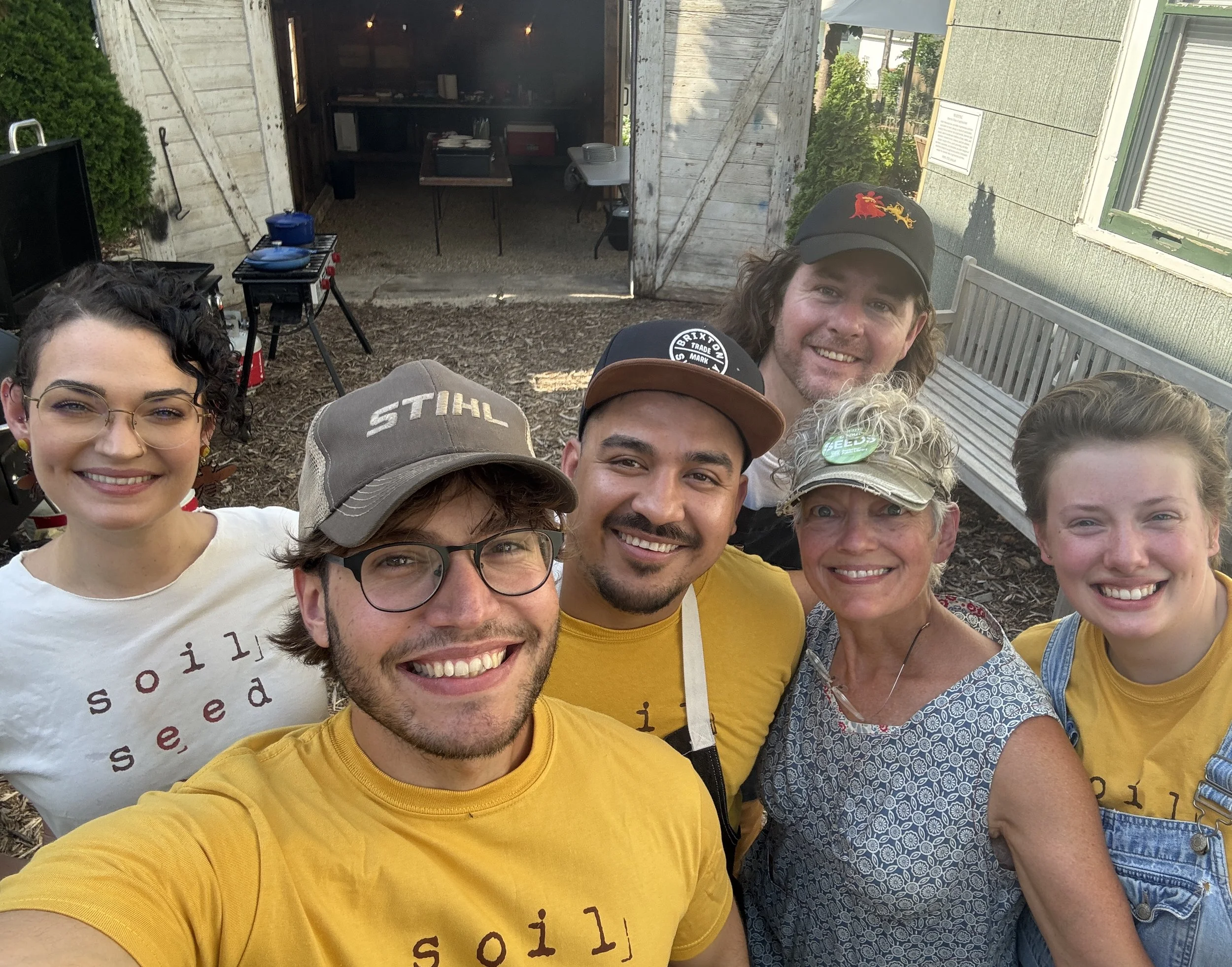 A group of people taking a selfie outdoors, some wearing yellow shirts, in front of a wooden shed with an open door and a small table inside. They are smiling and appear cheerful.