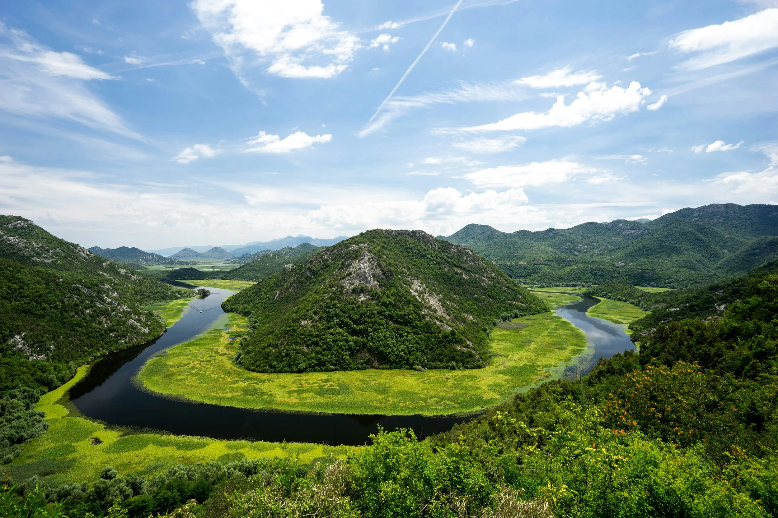 A scenic landscape of a winding river flowing through lush green hills and mountains under a partly cloudy sky.