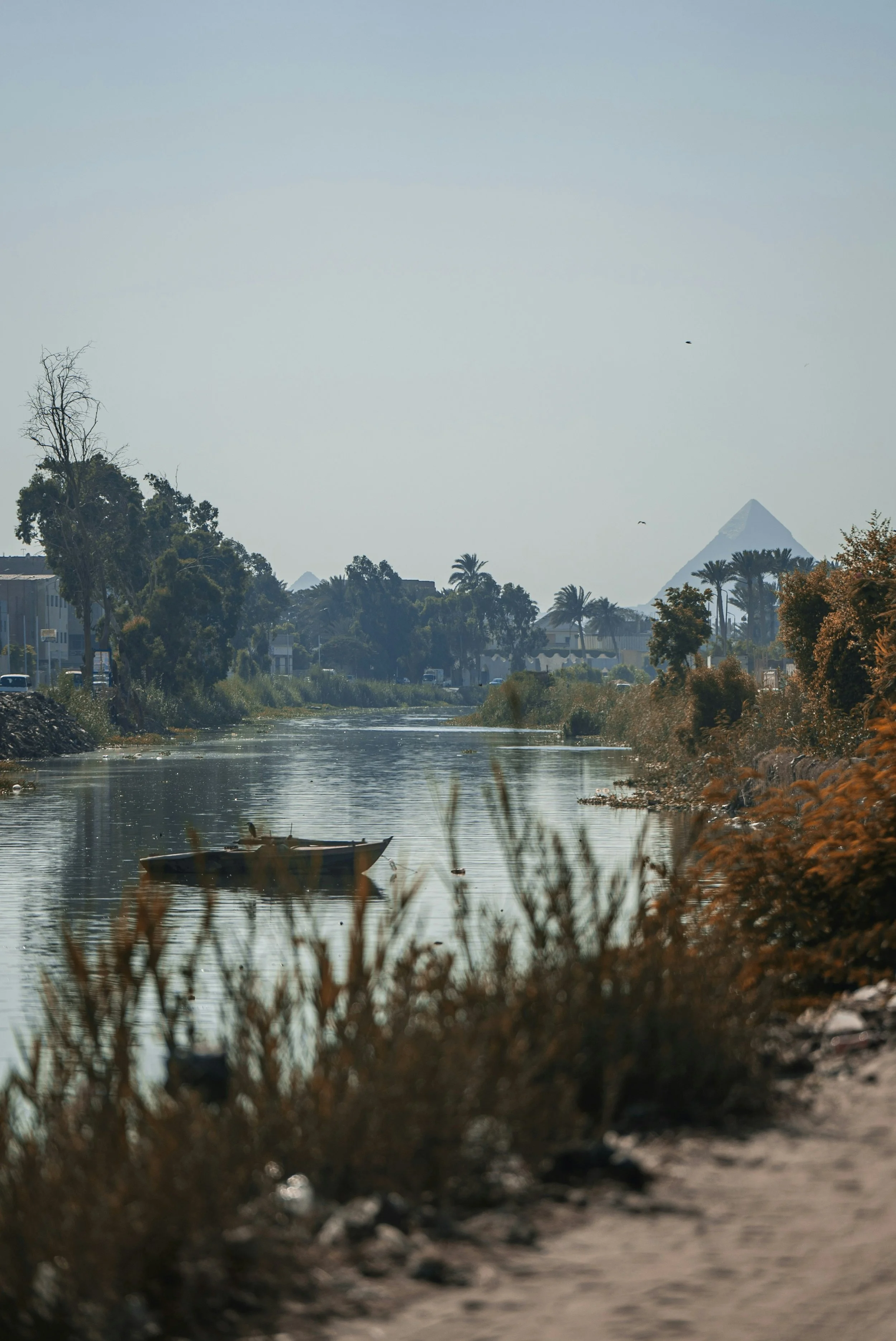 A river with a small boat floating on the water, with trees along the banks, and pyramids visible in the distance under a hazy sky.