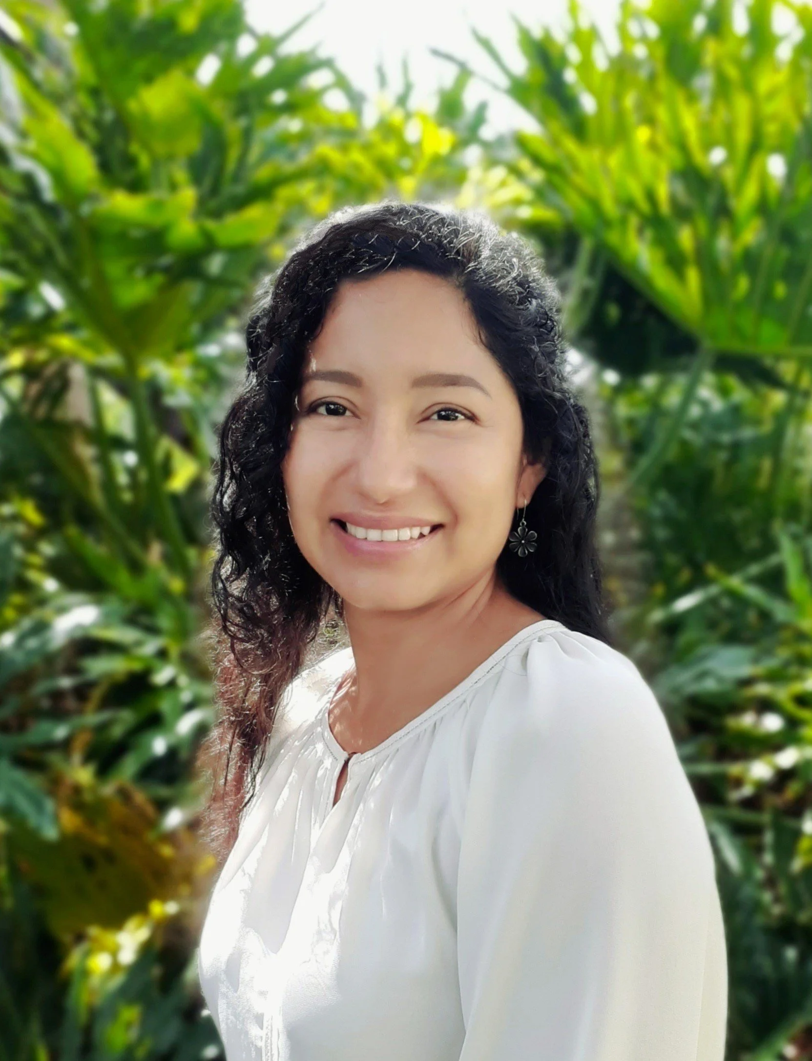 A woman smiling outdoors with green leafy plants in the background.