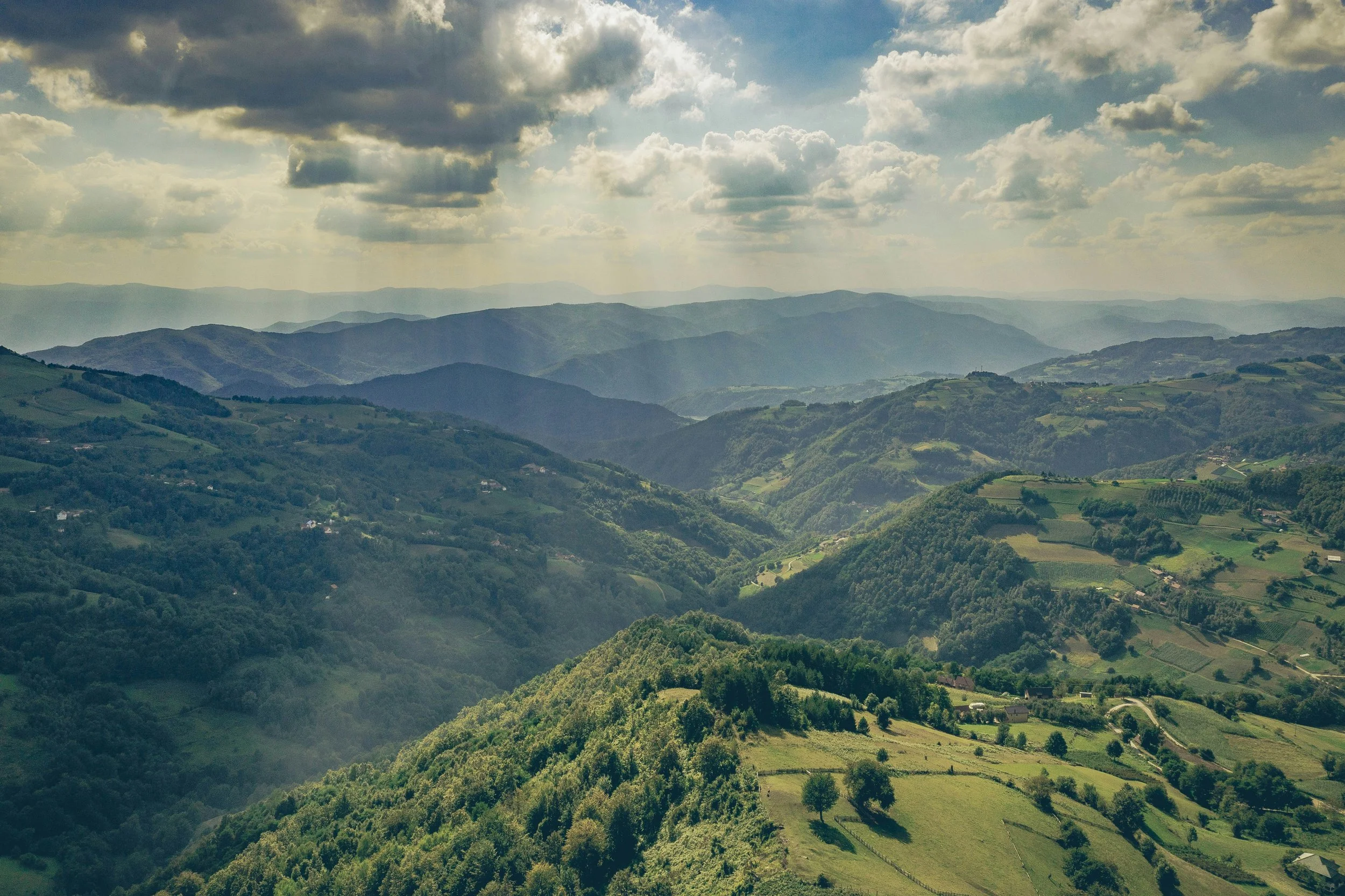 A scenic view of rolling green hills and mountains under a partly cloudy sky with sunlight breaking through the clouds.