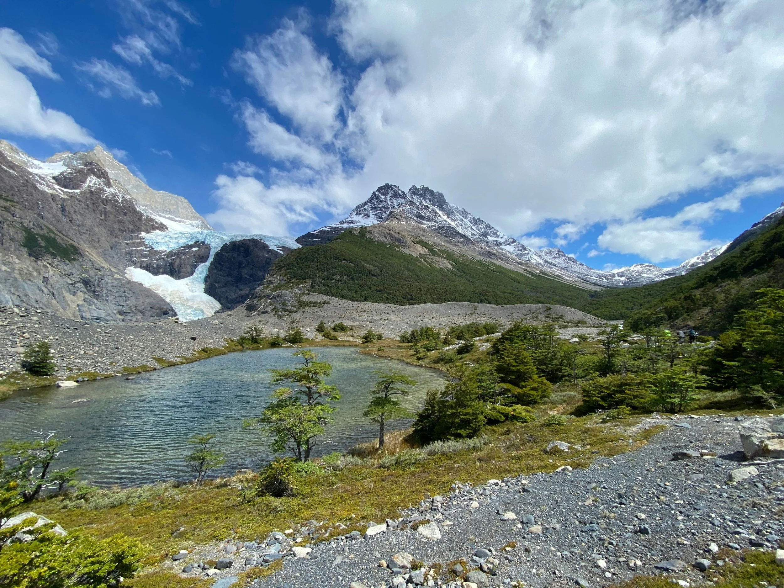 Mountain landscape with snow-capped peaks, a glacier, green vegetation, and a body of water in the foreground under a partly cloudy sky.
