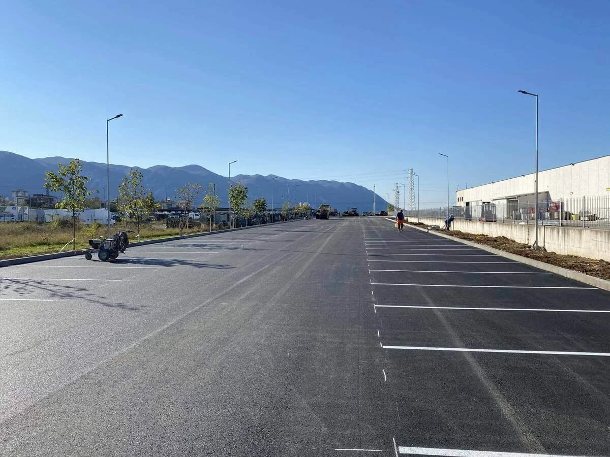 Mostly empty parking lot with a few people working, small trees, street lamps, and mountains in the background under a clear blue sky.