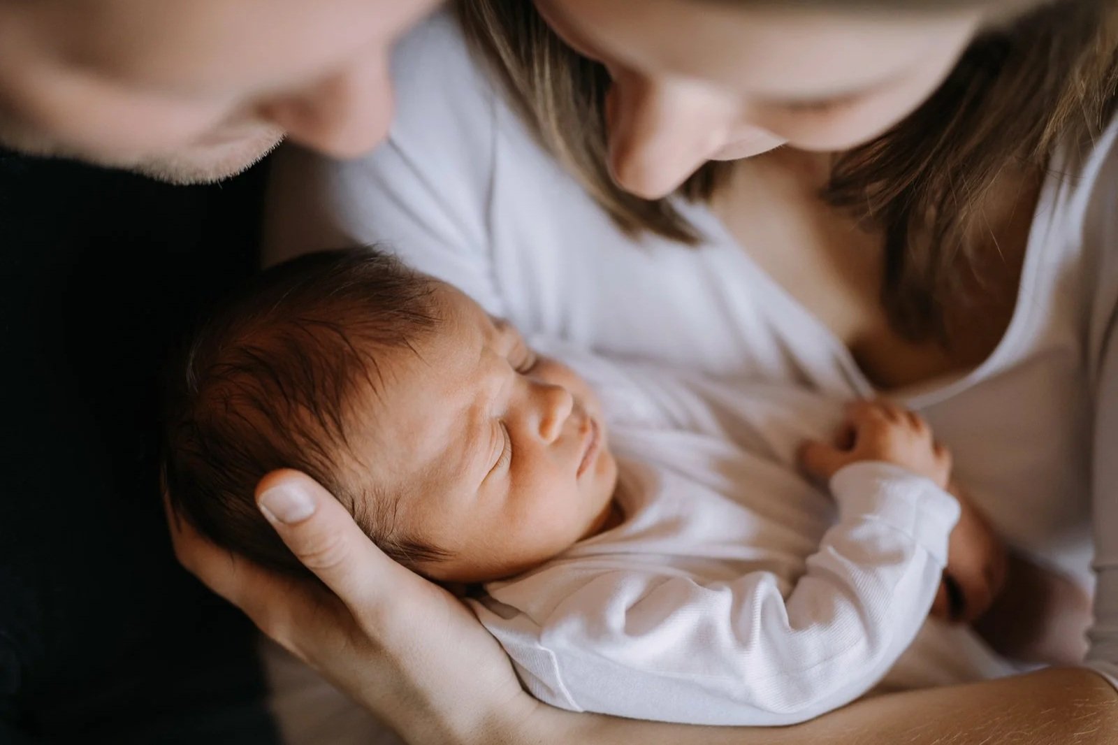 Ein Baby liegt im Arm eines Erwachsenen, während ein zweiter Erwachsener über dem Baby gebeugt ist. Das Baby schläft eingerollt mit geschlossenen Augen.