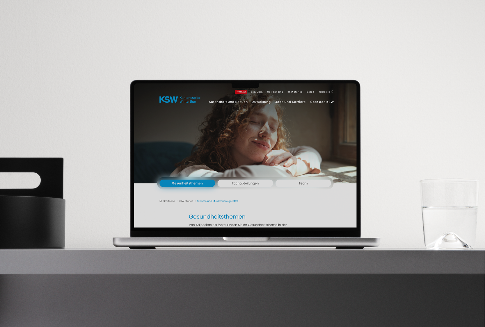 Laptop auf einem Tisch zeigt die Webseite des Kantonsspitals Winterthur mit einem Bild einer entspannten, lächelnden Frau, die die Hände unter dem Kopf liegen hat, bei Tageslicht. Rechts im Bild ein Glas Wasser, links eine schwarze Tasse.