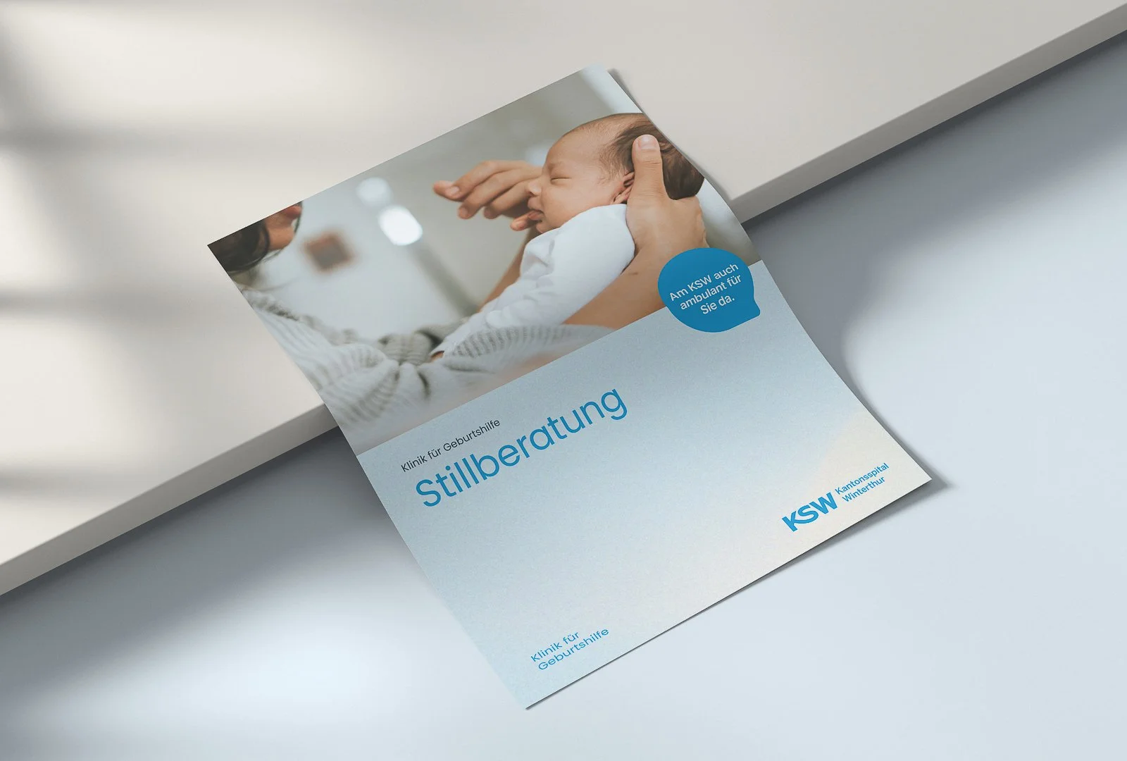 Flyer für Stillberatungen der Klinik für Geburtshilfe mit einem Bild eines Babys und einer Frau, die es hält, auf einem hellen Tisch.