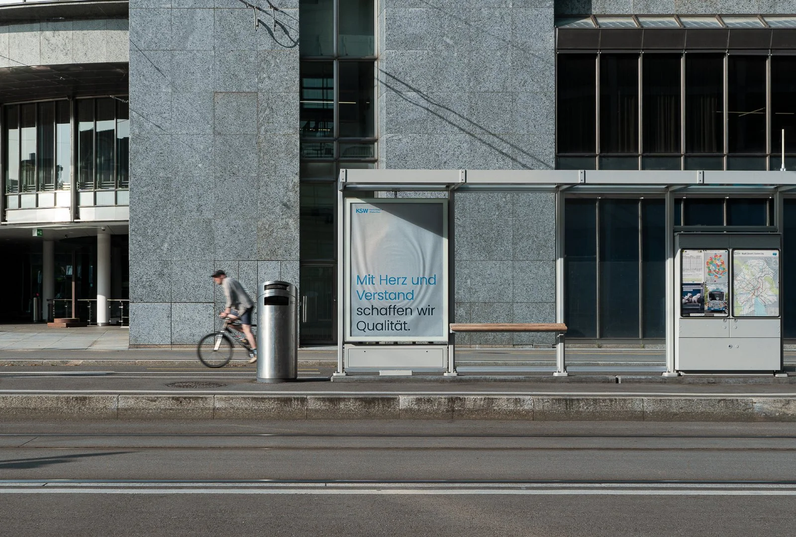 Bushaltestelle in einer Stadt mit einer digitalen Anzeigetafel, einem Poster mit einem Slogan, einem Zeitungs- und Stadtplan-Poster sowie einem Fahrradfahrer, der vorbeifährt.