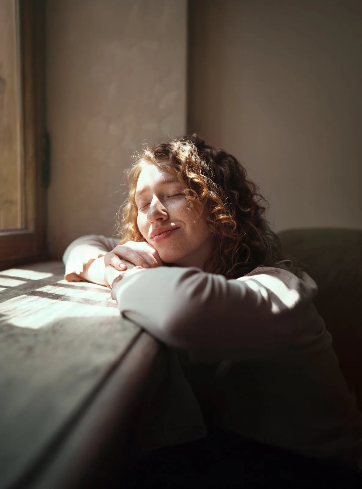 Eine lächelnde junge Frau mit lockigem roten Haar, die entspannt am Fenster sitzt und die Augen geschlossen hat, während sie das Sonnenlicht genießt.