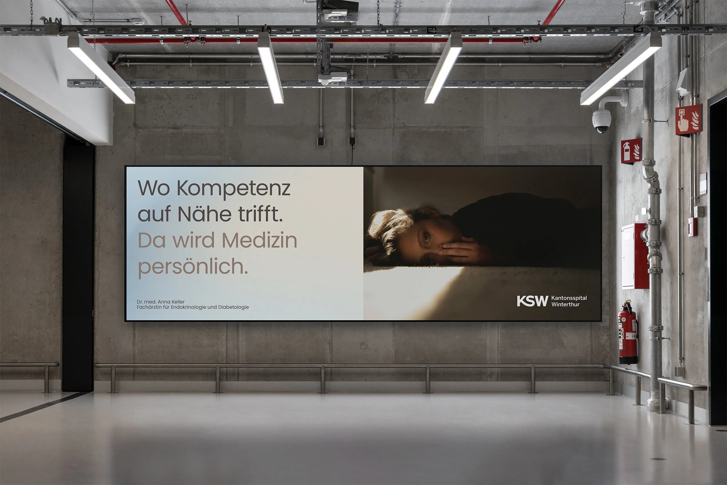 Ein Wartebereich in einem Krankenhaus mit einem großen Bildschirm, auf dem ein medizinischer Vortrag angezeigt wird. Der Bildschirm zeigt einen Text in Deutsch mit dem Titel "Wo Kompetenz auf Nähe trifft. Da wird Medizin persönlich" und das Logo des 