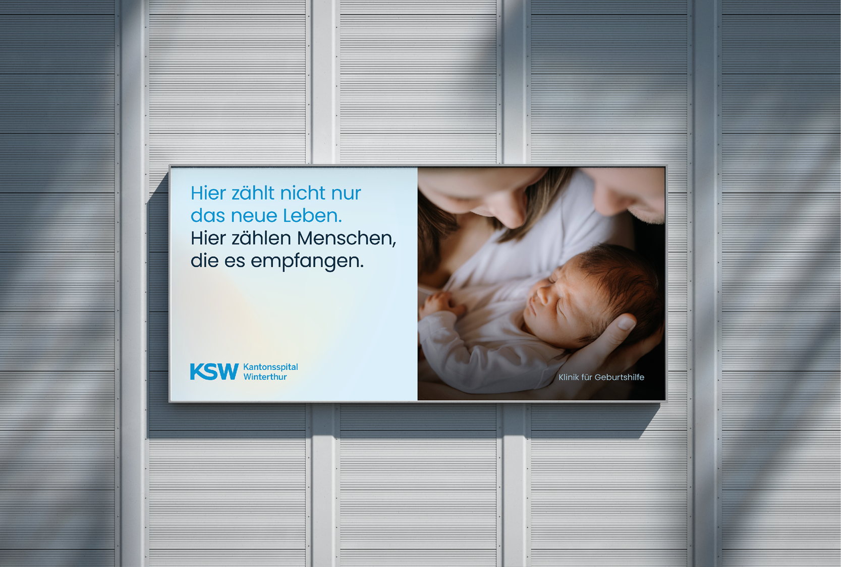 Ein leeres Werbeschild mit einem Foto eines Neugeborenen, das von einer Frau gehalten wird, und einem Text auf der linken Seite, der auf Deutsch lautet: "Hier zählt nicht nur das neue Leben. Hier zählen Menschen, die es empfangen." Das Schild ist an 