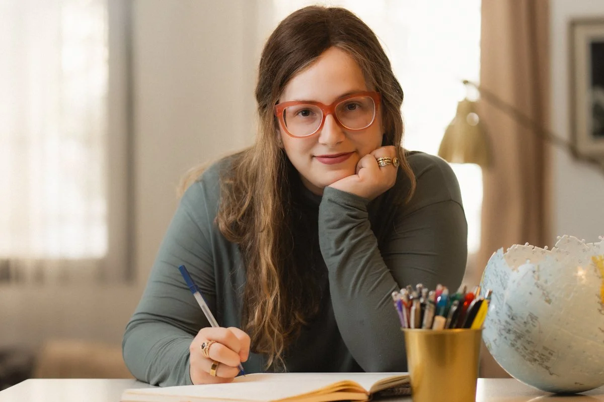 A woman with long brown hair, red glasses, and wearing a gray long sleeve top, sitting at a table with a world globe, a container of colored pencils, and an open notebook, resting her head on her hand and smiling at the camera.