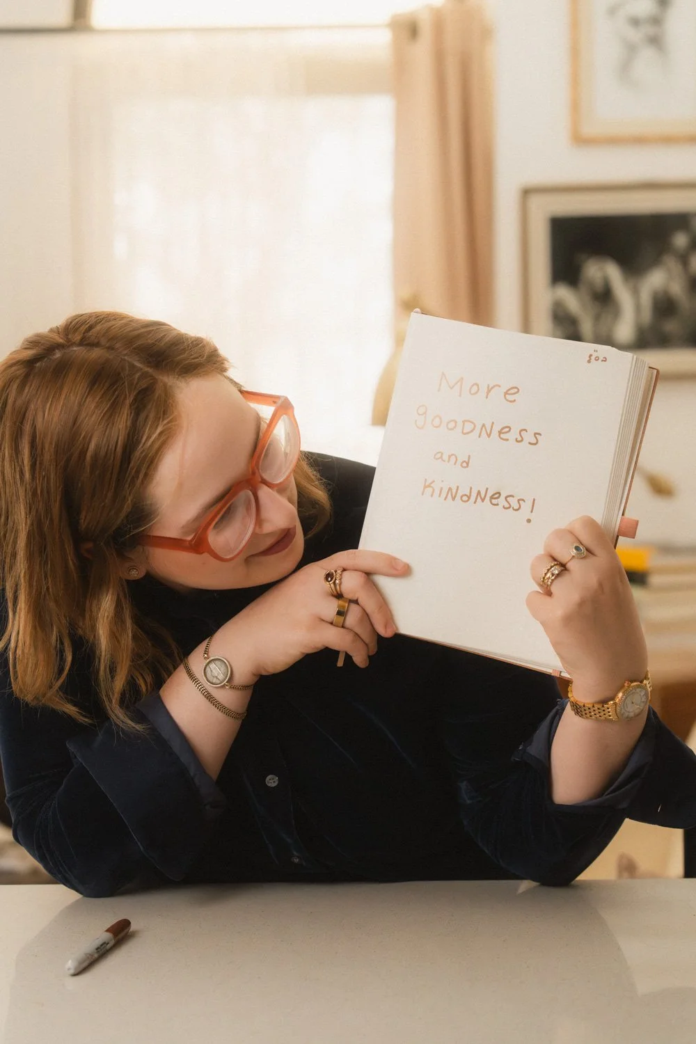 A woman with red hair and glasses holding a notebook that says 'More GOODNESS and KINDNESS!' inside a cozy, well-lit room.