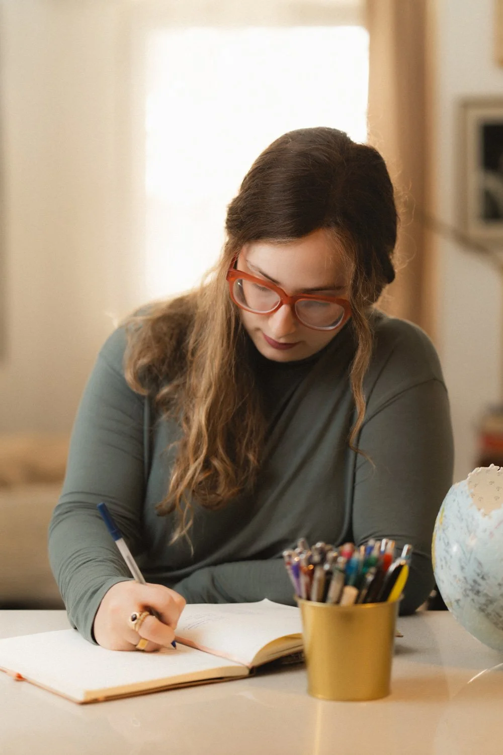 A woman with long wavy hair and orange glasses writing in a notebook at a desk, with a yellow cup of colored pens and a globe nearby.