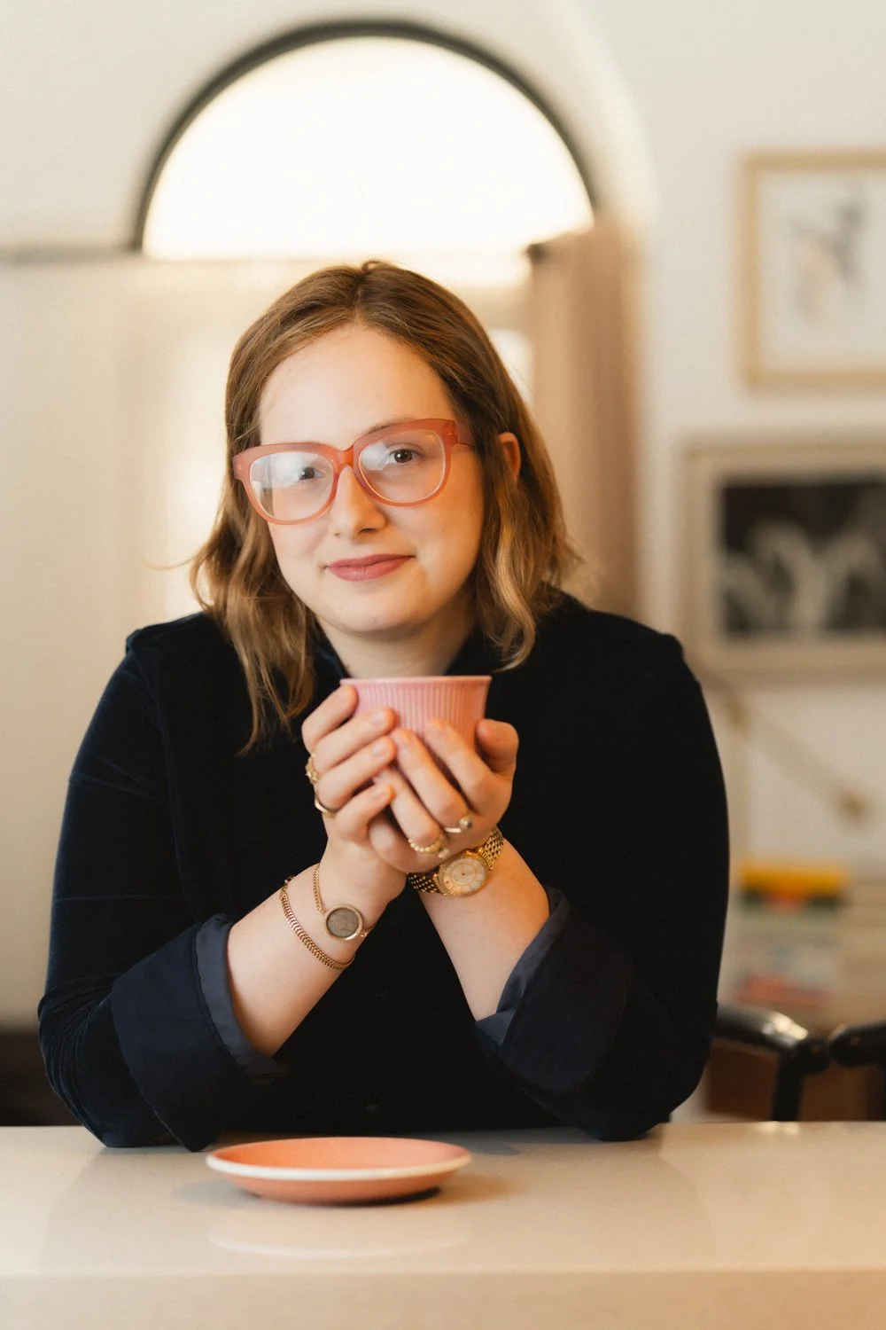 A woman with shoulder-length light brown hair, wearing large pink glasses, holds a pink teacup with both hands, smiling gently. She wears a black long-sleeve top, gold and silver jewelry, and a gold wristwatch. A matching pink saucer is on the white table in front of her. The background shows a softly lit room with framed pictures on the wall.