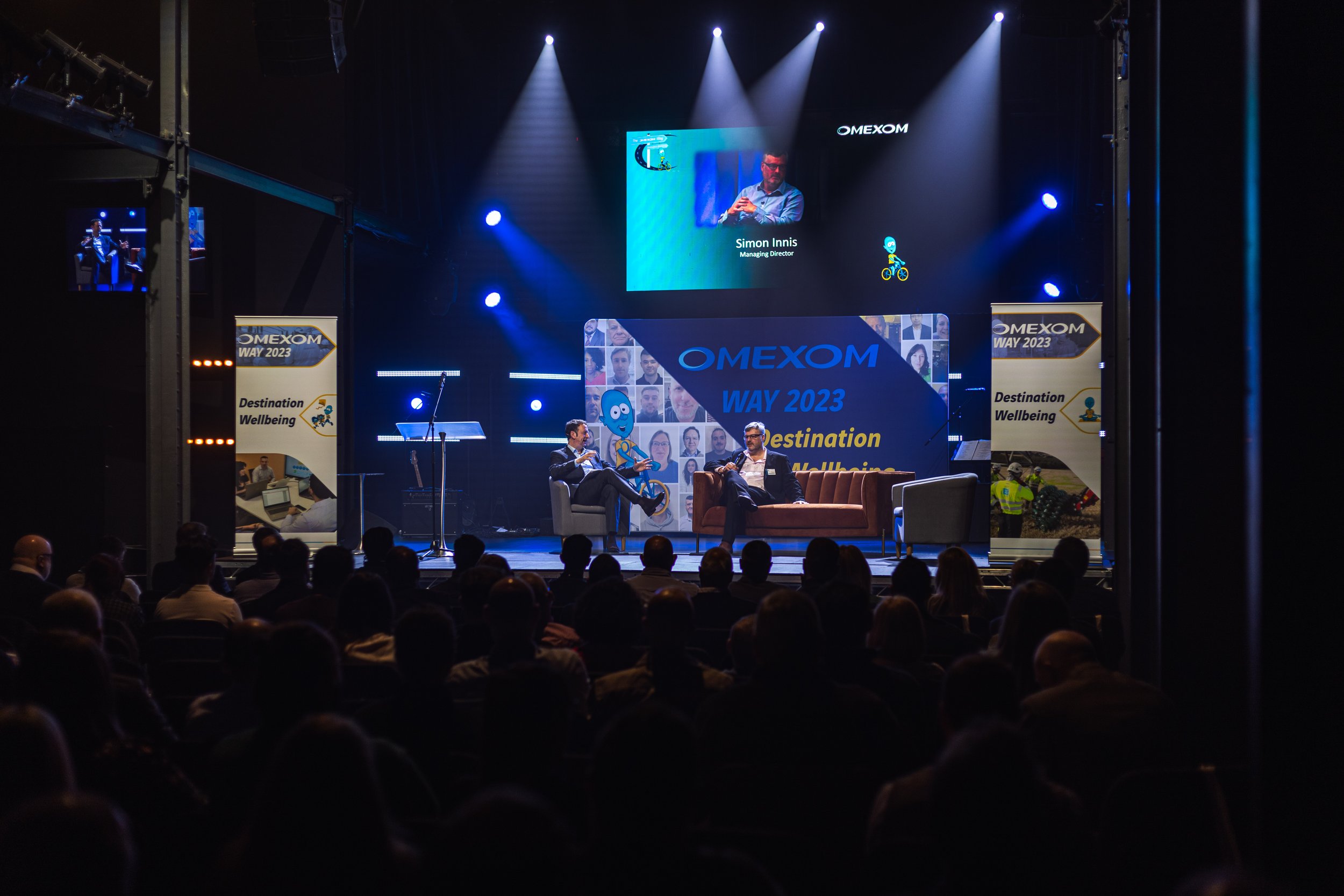 A conference event live on stage with two speakers talking in front of a large LED screen, an audience seated in front, and stage lighting.