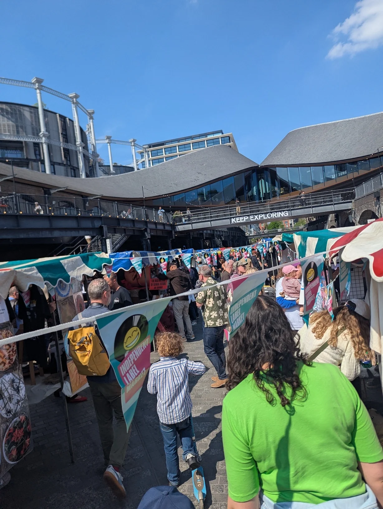 Crowds browsing stalls at the Independent Label Market at Coal Drops Yard, Kings Cross, London on a sunny day, with Independent Label Market bunting in the foreground and an eight year old on a scooter